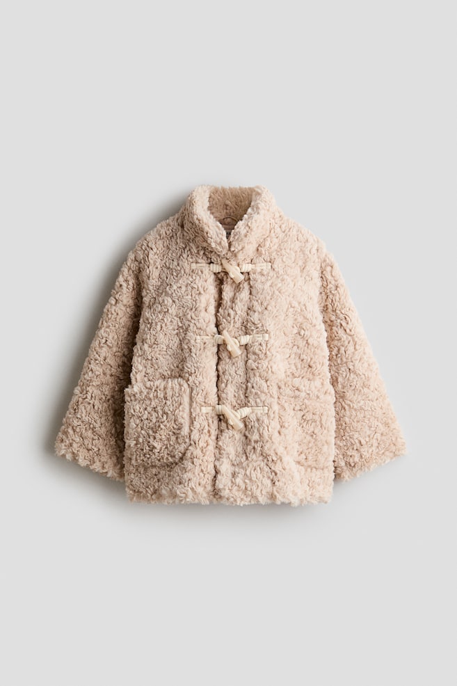 En ljusbeige, mjuk duffelrock i teddypÃ¤lsfleece visas framifrÃ¥n och framhÃ¤ver sitt strukturerade lockiga material, sin hÃ¶ga stÃ¥krage och lÃ¥nga Ã¤rmar. Den har tre krÃ¤mfÃ¤rgade toggle-knÃ¤ppningar med repÃ¶glor lÃ¤ngs mitten samt tvÃ¥ matchande utanpÃ¥fickor framtill. - 1