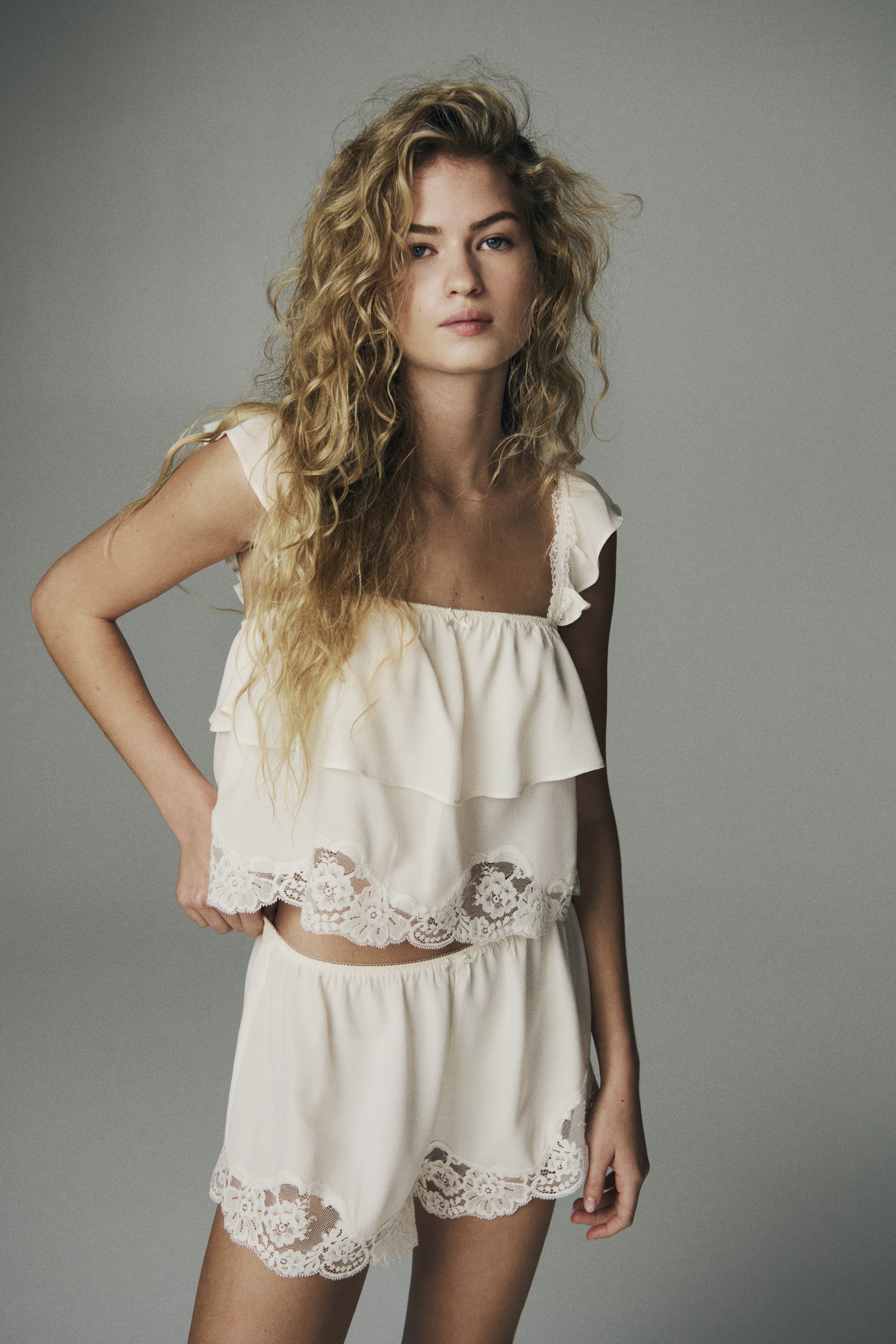 Büyük resmi görüntüle: Model, krem renkli dantel detaylı bir pijama üstü giyiyor; kare yakalı, fırfırlı kap kollu ve alt kenarında ince işlemeli çiçek danteli bulunan katmanlı fırfır detaylara sahip. Model ayrıca uyumlu dantel kenarlı pijama şortu giyiyor ve üstün etek ucunu hafifçe düzeltiyor.