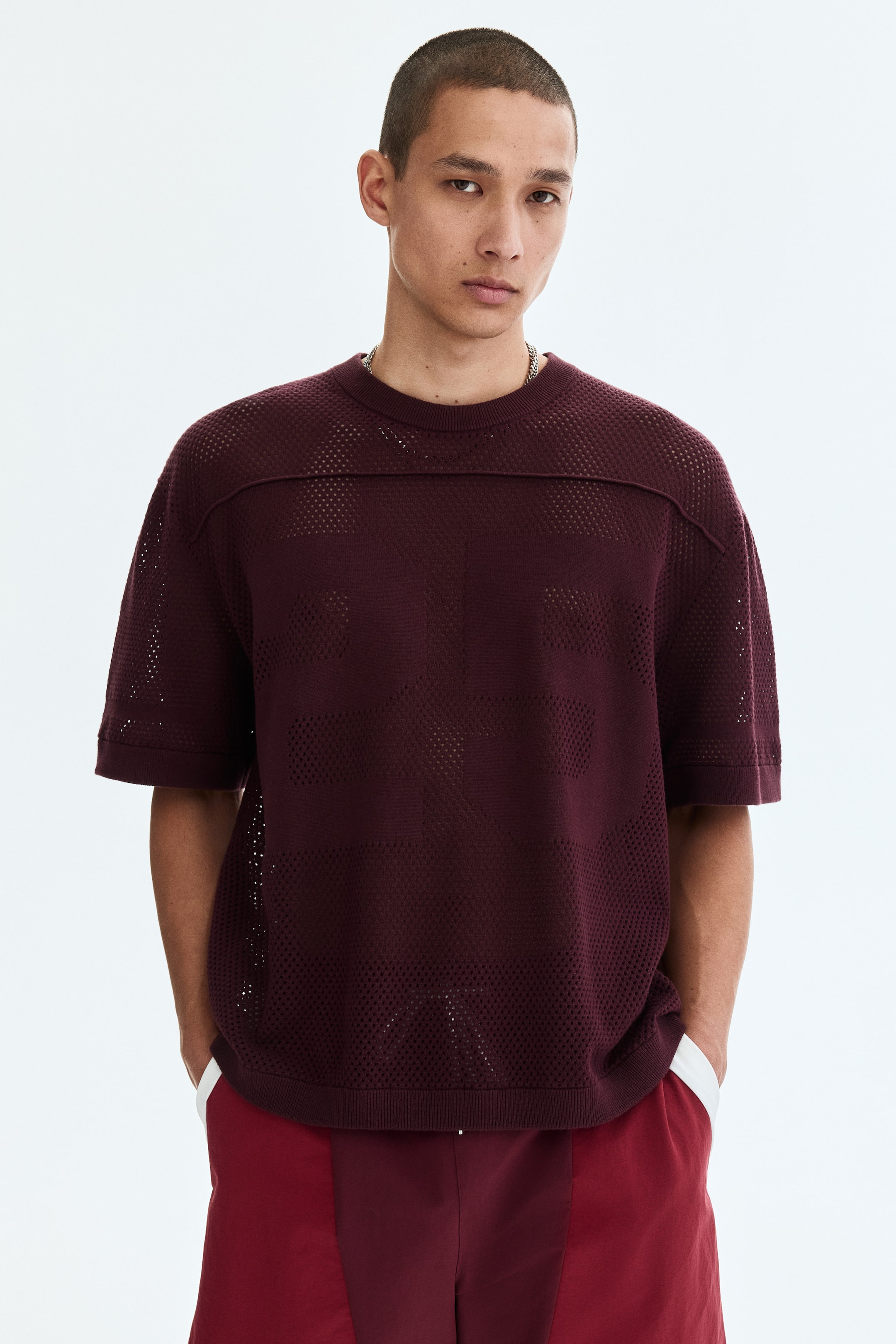 View larger image: The model is wearing a burgundy boxy-style T-shirt crafted from hole-knit cotton, featuring a round ribbed neckline, piping along the front yoke, and ribbed cuffs and hem. The model stands with his hands in the pockets of burgundy shorts.