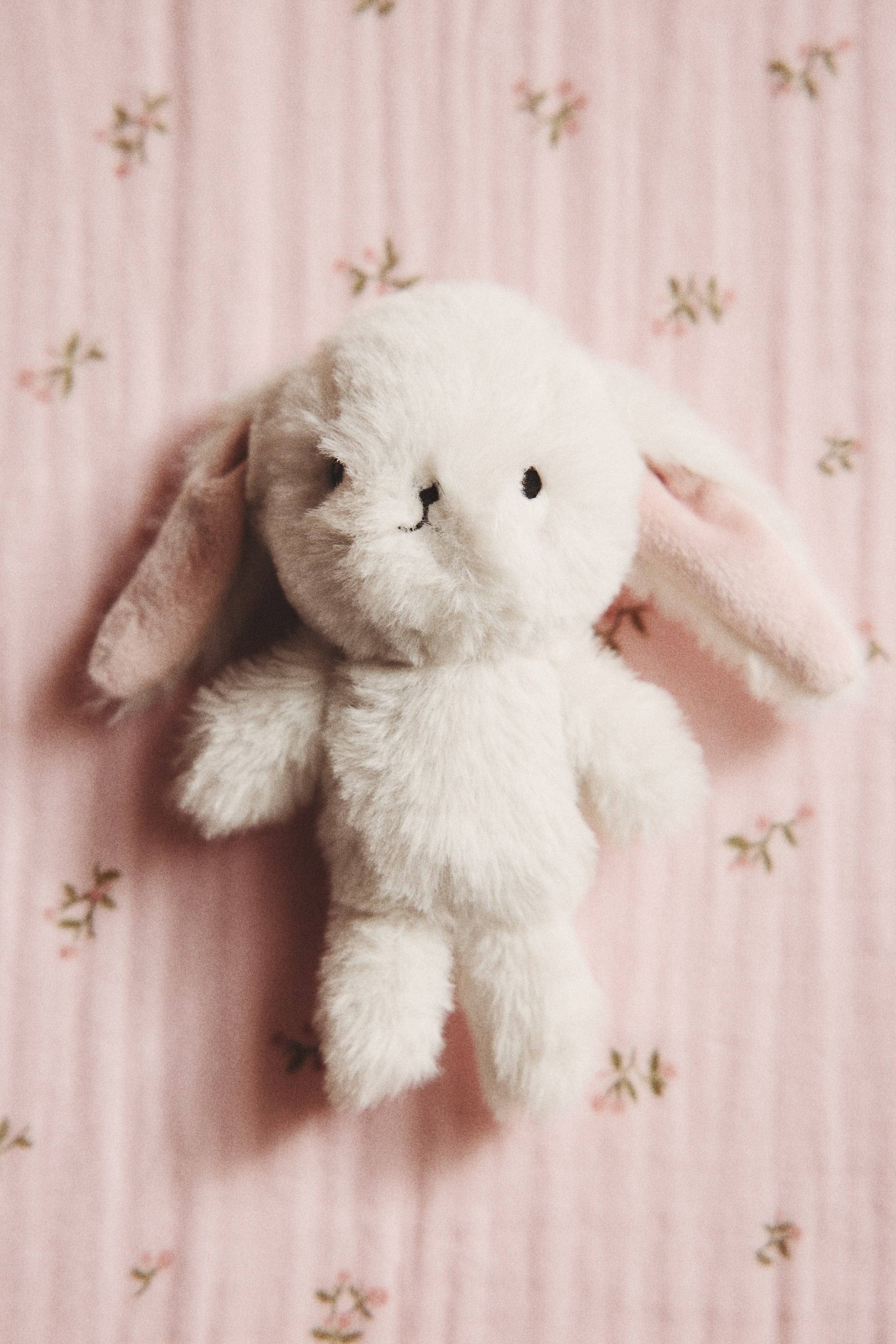View larger image: A padded white bunny-shaped baby rattle is facing front, featuring a soft pile texture and embroidered black eyes, nose, and mouth. The long ears have pink inner surfaces.
