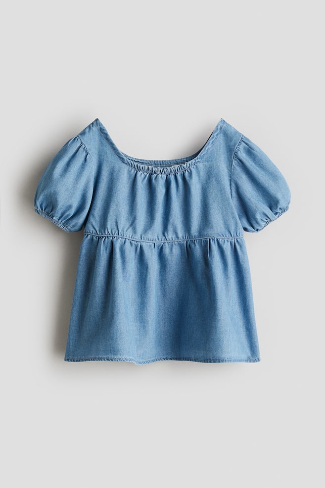 A light blue, oversized blouse in a soft weave is showing as FRONT, featuring a square, elasticated neckline and short puff sleeves with elasticated cuffs. It is designed with narrow elastication above the waist and a flared hem. - 1