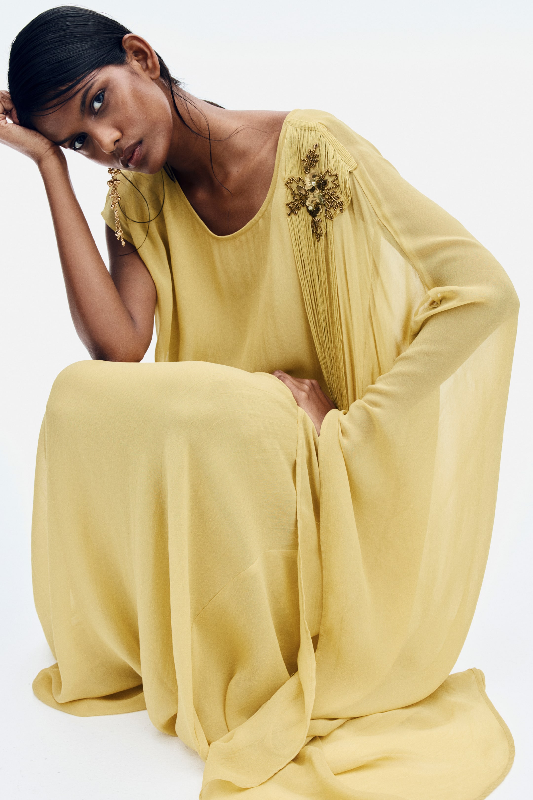 View larger image: A long, loose-fitting yellow dress in a lightweight fabric is worn by the model who is crouching. The asymmetrical dress features a sleeveless side and a long, flowing cape sleeve on the other, decorated with a beaded floral embellishment and long hanging fringes on the shoulder.