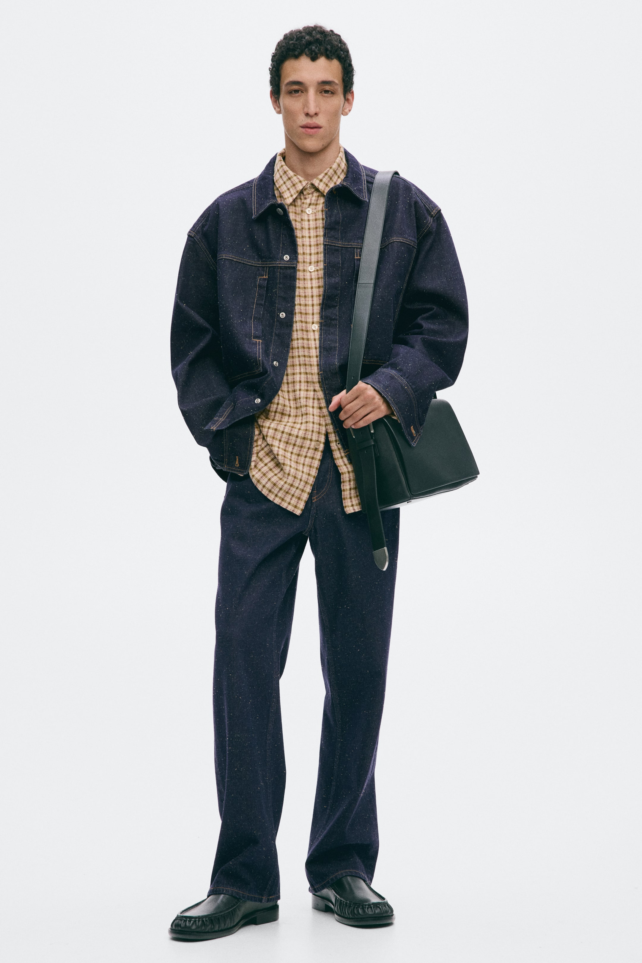 Grösseres Bild ansehen: Ein Overshirt aus festem Baumwoll-Denim wird vom Model getragen und verfügt über einen Kragen, eine Knopfleiste vorn, aufgesetzte Brusttaschen, lange Ärmel mit leicht überschnittenen Schultern, Ärmelschlitze und geknöpfte Manschetten. Es wird über einem karierten Hemd getragen, und die rechte Hand des Models ruht in einer Tasche.