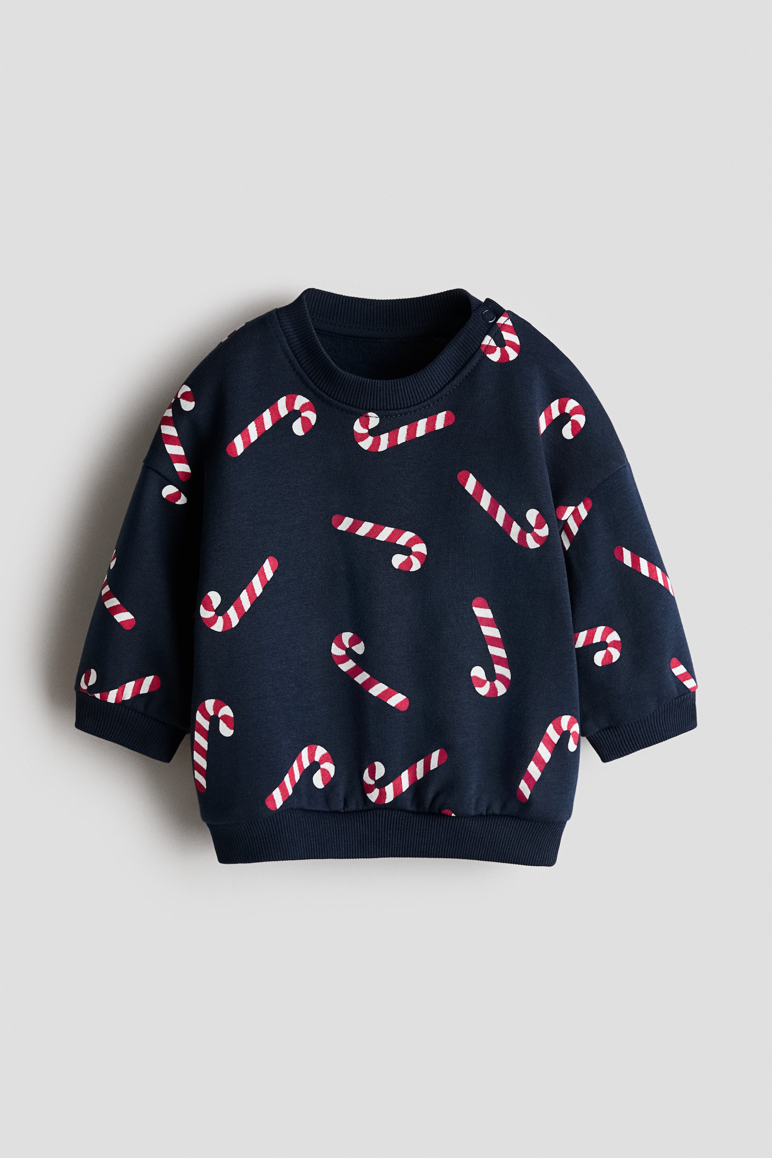 Ein marineblauer Pullover mit Print ist von vorne zu sehen und zeigt ein Allover-Muster aus rot-weiÃ gestreiften Zuckerstangen. Das KleidungsstÃ¼ck verfÃ¼gt Ã¼ber einen gerippten Rundhalsausschnitt mit DruckknÃ¶pfen an der Schulter, gerippte BÃ¼ndchen und einen gerippten Saum.