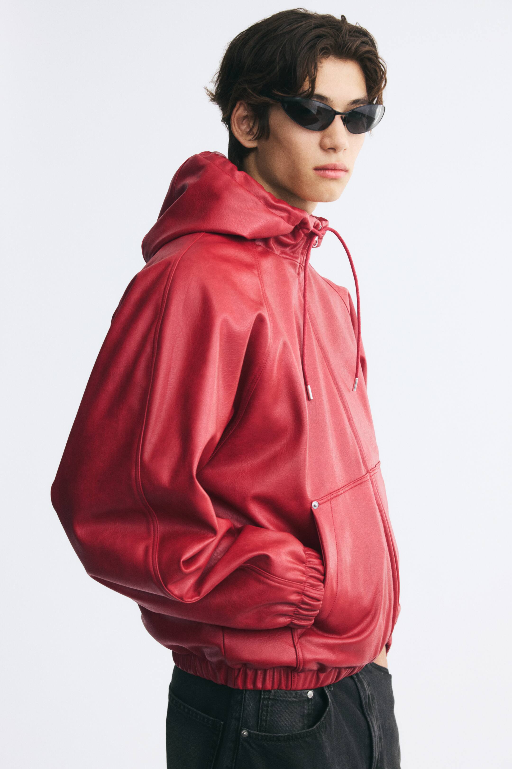 Agrandir l'image: Une veste rouge en tissu enduit, dotée d’une capuche à cordon de serrage ainsi que de poignets et d’un ourlet élastiqués, est portée par le mannequin. Le mannequin est debout, une main glissée dans la poche avant ouverte de la veste, qui est assortie à un jean noir et à des lunettes de soleil.