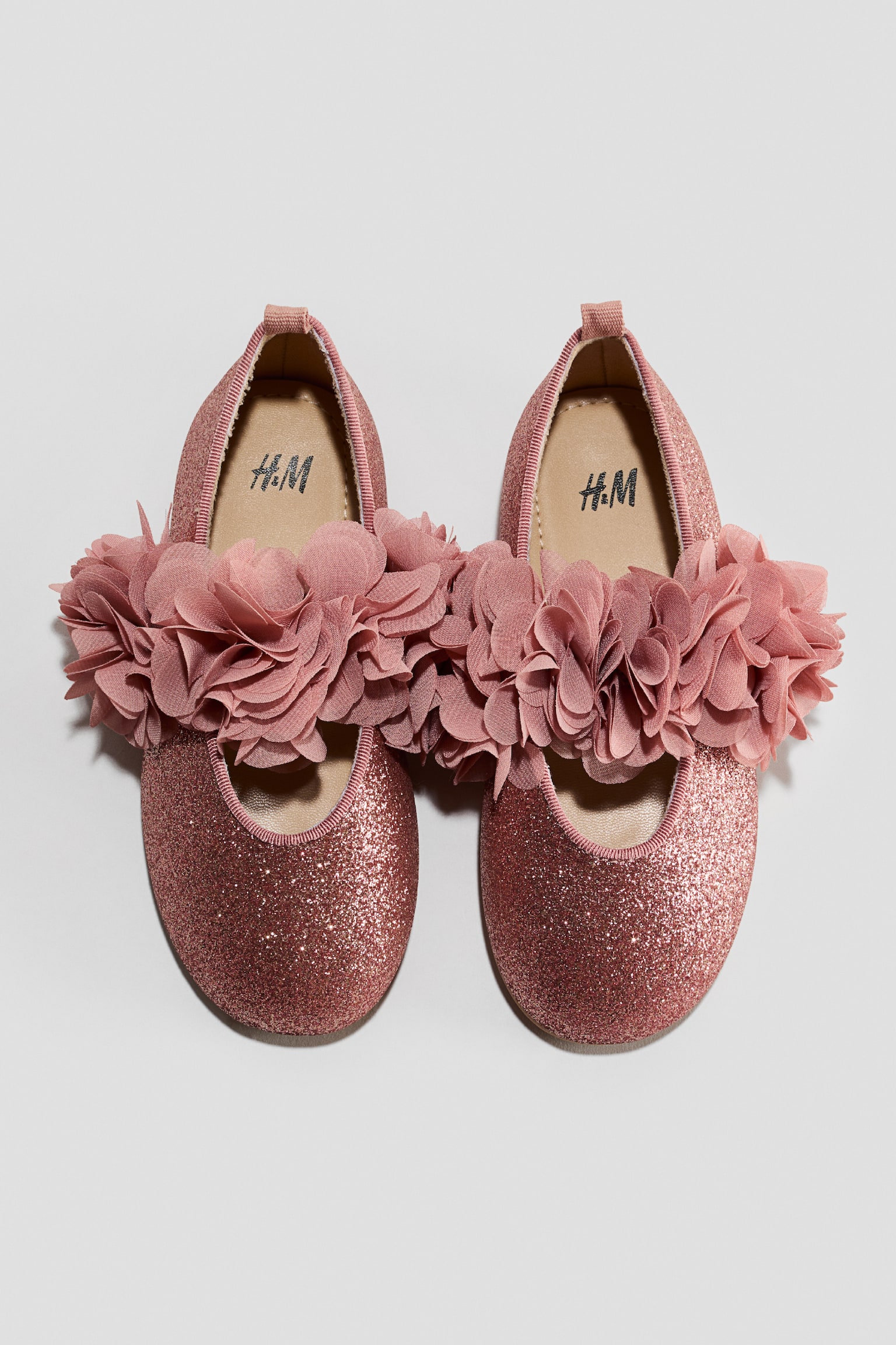 A pair of pink glittery ballet flats is shown front-facing in a top-down view, featuring a sparkling pink upper and a large, ruffled fabric flower embellishment across the instep. The shoes are designed with rounded toes, a beige inner lining, and small pull tabs at the heel.