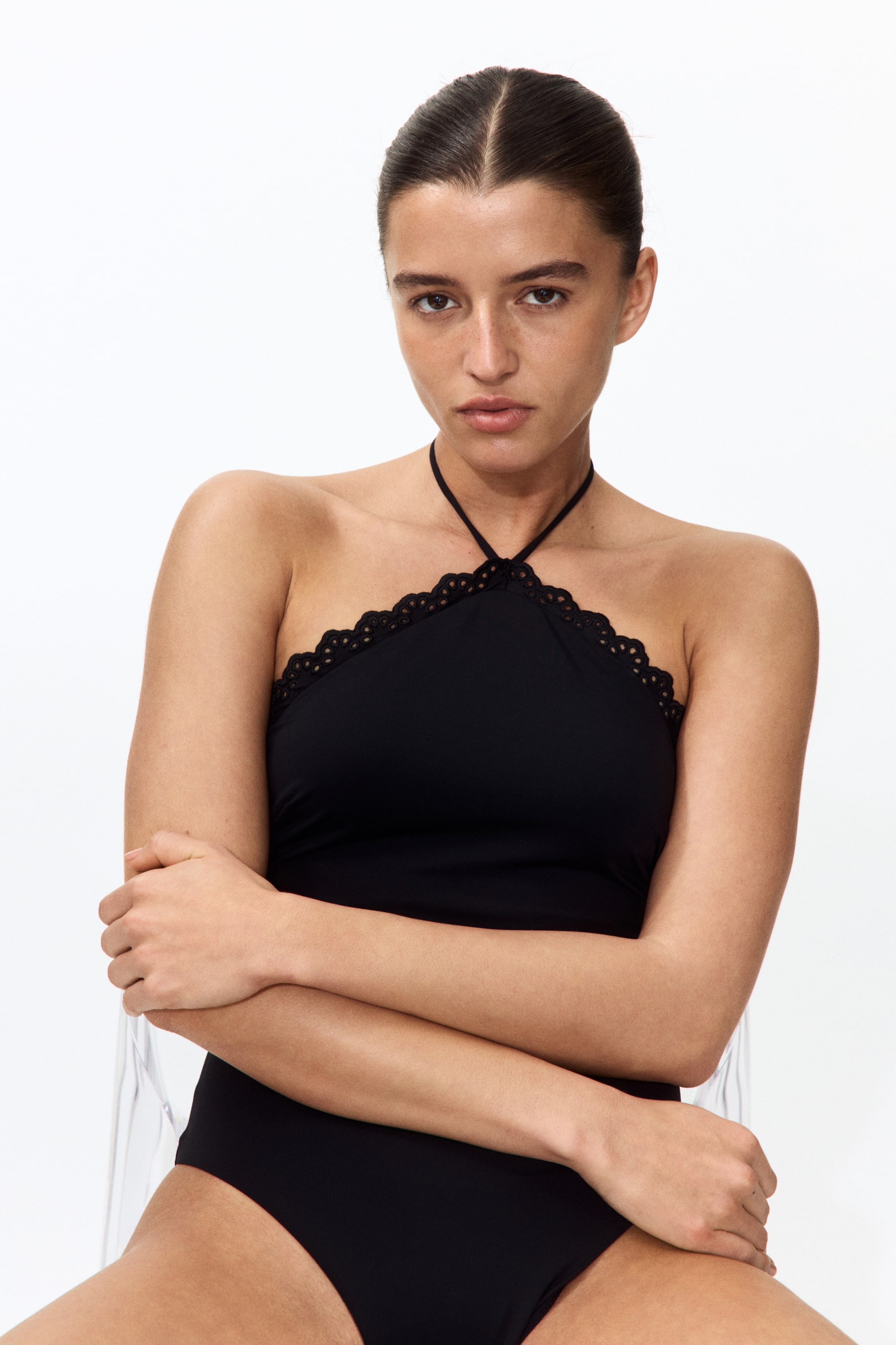 View larger image: The model is wearing a black swimsuit with a halterneck design, featuring narrow ties. The top edge is adorned with a scalloped trim and broderie anglaise detailing.