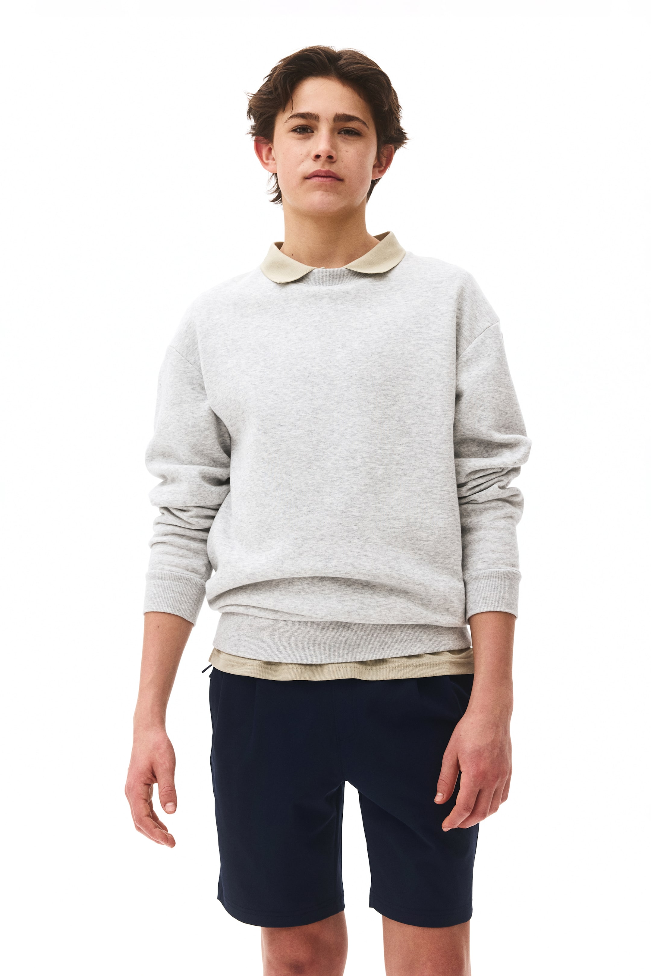 View larger image: A light gray crew neck sweatshirt is shown from the front, featuring long sleeves with ribbed cuffs and a ribbed hem. A light beige collared shirt is layered underneath, with its collar visible above the sweatshirt's neckline and its hem extending below.