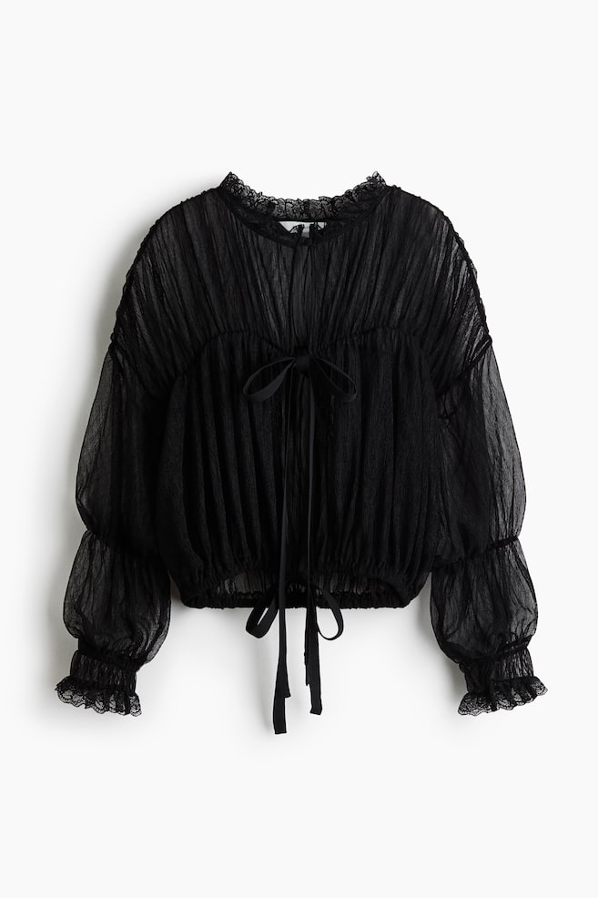 Un chemisier noir en jersey froissé transparent est présenté de face, avec un col rond bordé de volants, des épaules tombantes et des manches longues à poignets froncés bordés de volants. Le chemisier met en valeur des coutures froncées sur lâensemble, une ouverture sur le devant fermée par un fin nÅud à nouer, ainsi quâun bas resserrable avec liens pendants qui créent une silhouette volumineuse. - 1