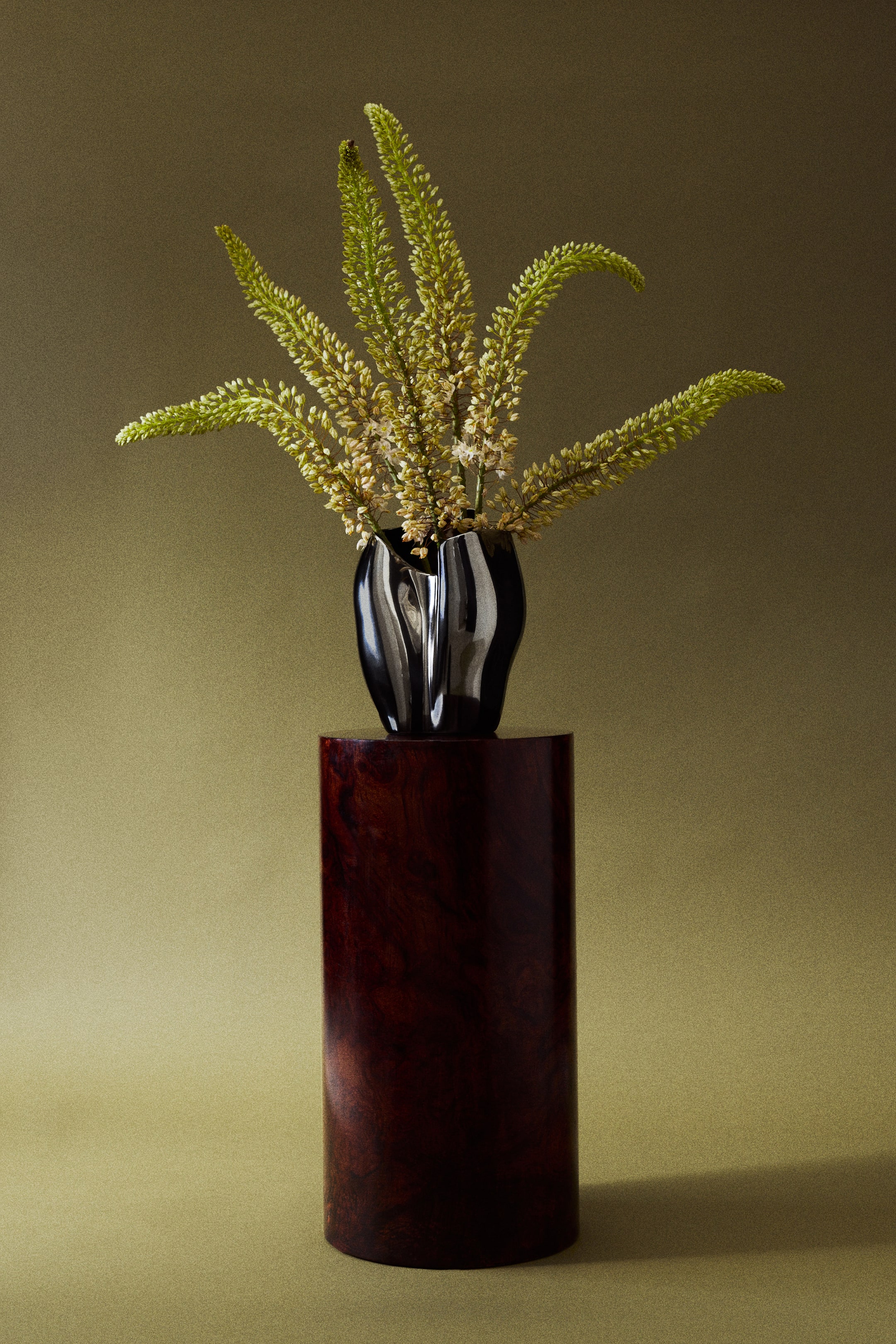 View larger image: A large stoneware vase with a reflective, electroplated metallic finish and an asymmetrical, sculptural design stands facing forward on a dark wooden pedestal, holding several long, light green flowering stems branching outward.