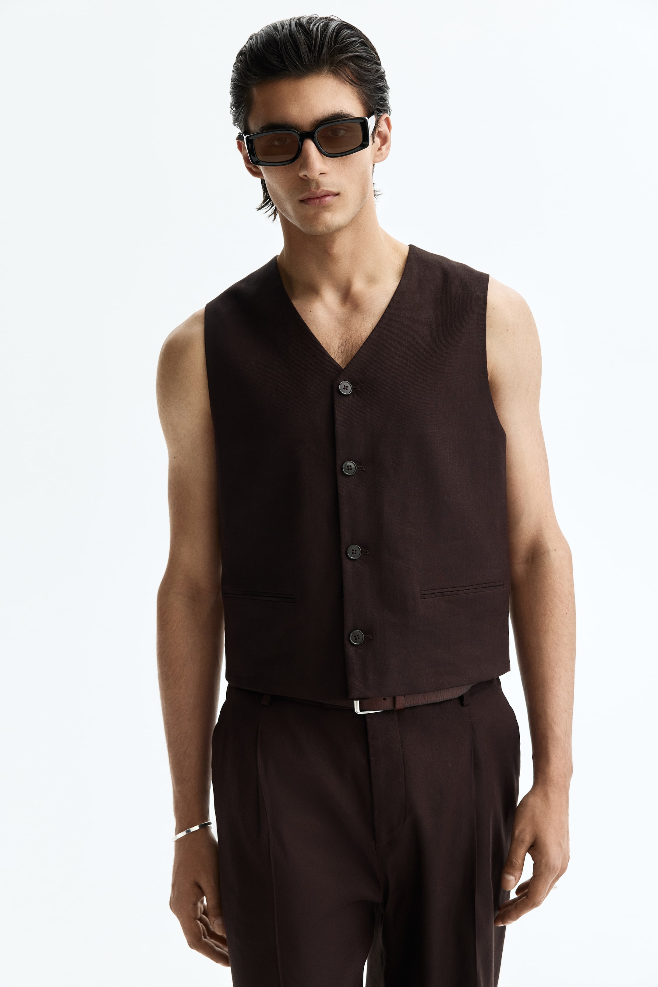 Visualizza immagine più grande: Un gilet marrone scuro e pantaloni coordinati sono presentati da un'angolazione frontale. Il gilet presenta uno scollo a V, una chiusura frontale con quattro bottoni scuri e due tasche a filetto orizzontali, mentre i pantaloni mostrano un davanti plissettato e una cintura marrone con una fibbia rettangolare chiara.
