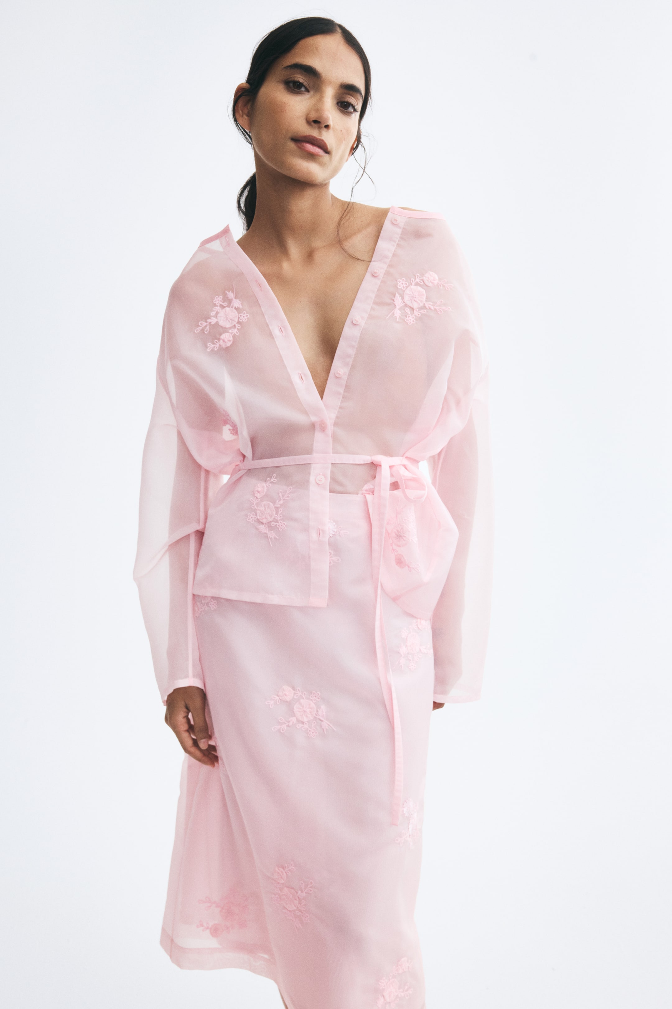 View larger image: The model is wearing a light pink embroidered organza skirt featuring a semi-sheer fabric detailed with tonal floral embroidery. The skirt is styled with a matching long-sleeved sheer organza shirt with a tied waist.