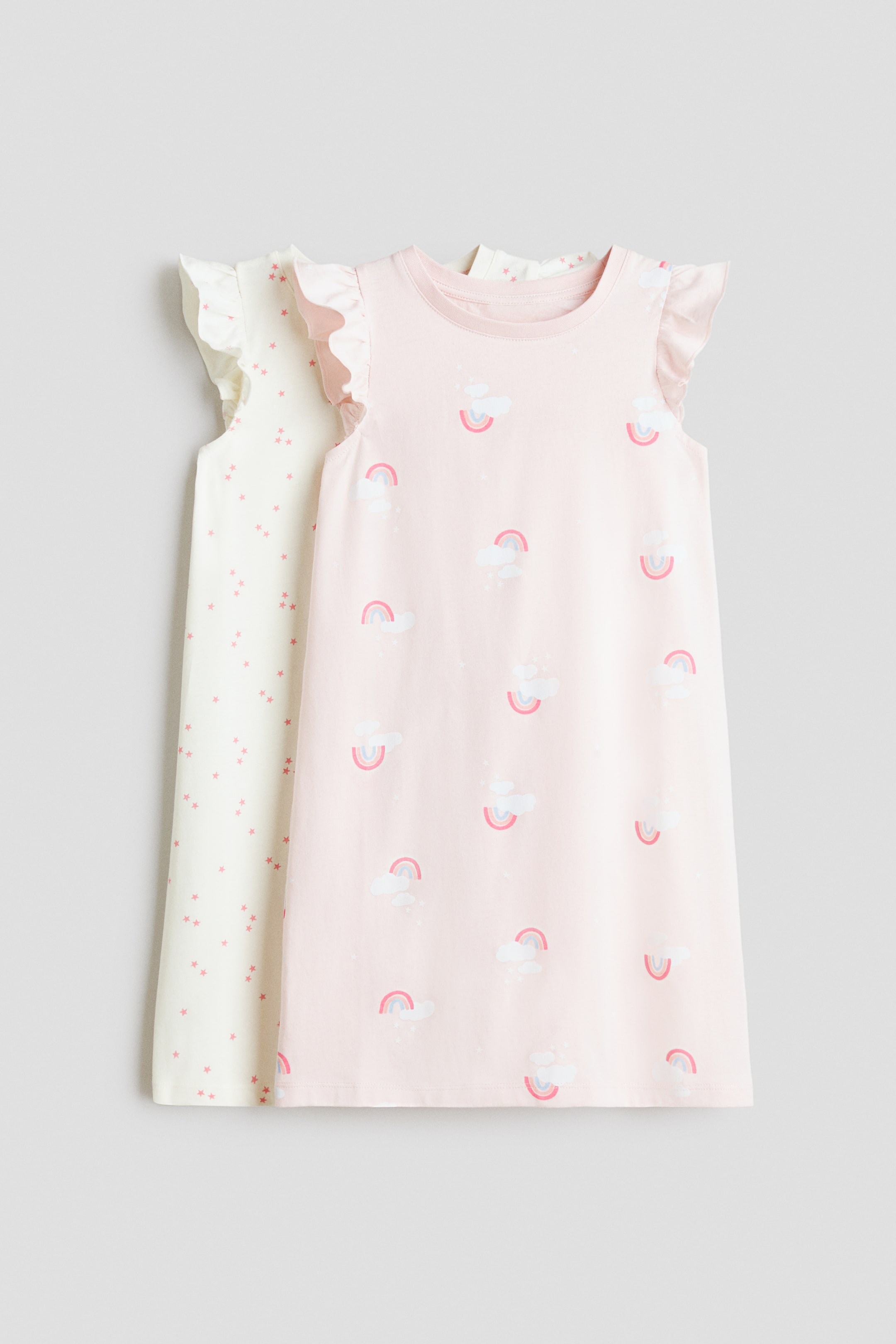 Ingrandisci l'immagine: Le camicie da notte sono realizzate in morbido jersey di cotone, con scollo rotondo e maniche a volant. Sono presentate frontalmente, con una camicia da notte in rosa chiaro con un motivo di nuvole bianche e piccoli arcobaleni, e l'altra in bianco sporco con stelle rosa sparse.