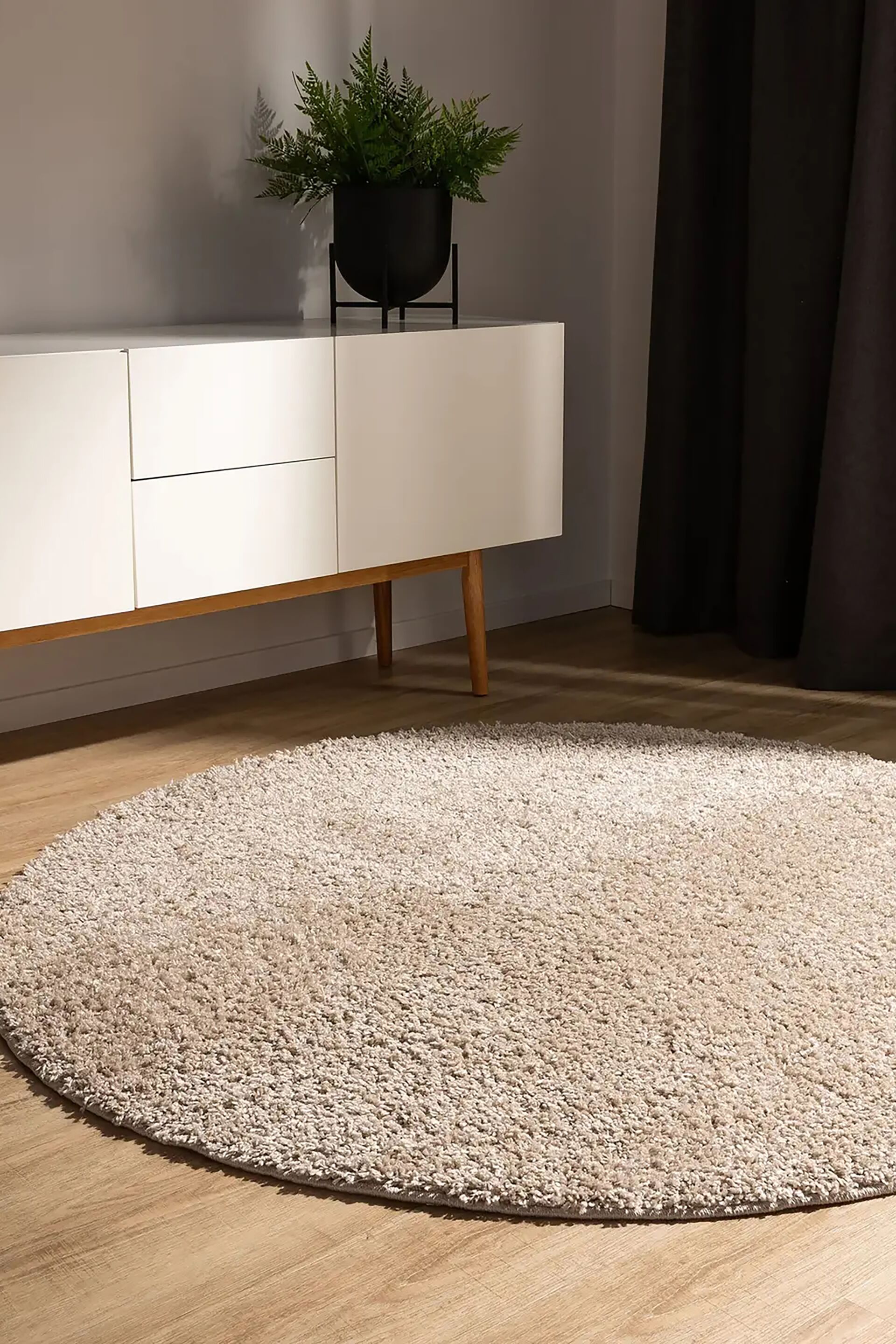 Um tapete shag redondo apresenta uma superfície macia e texturada numa cor taupe neutra. O tapete está estendido na horizontal, visto de cima, para realçar a sua forma circular e o pelo denso.