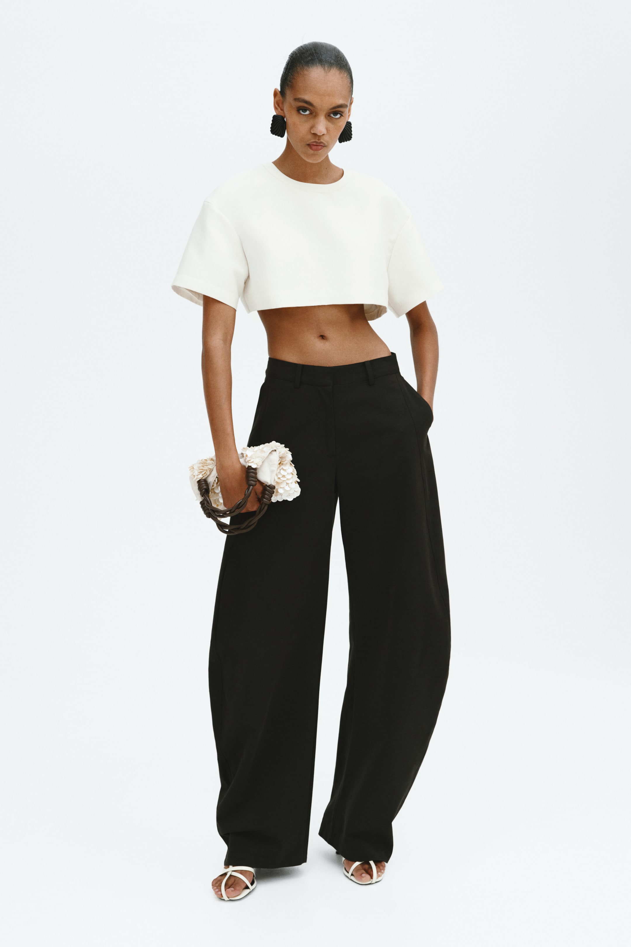 View larger image: Black wide-leg trousers feature a high waist, a flat front, and visible side pockets, with one hand casually placed in a pocket. These full-length trousers drape loosely to the ground, slightly pooling at the ankles and partially covering the feet, where white sandal straps can be seen.