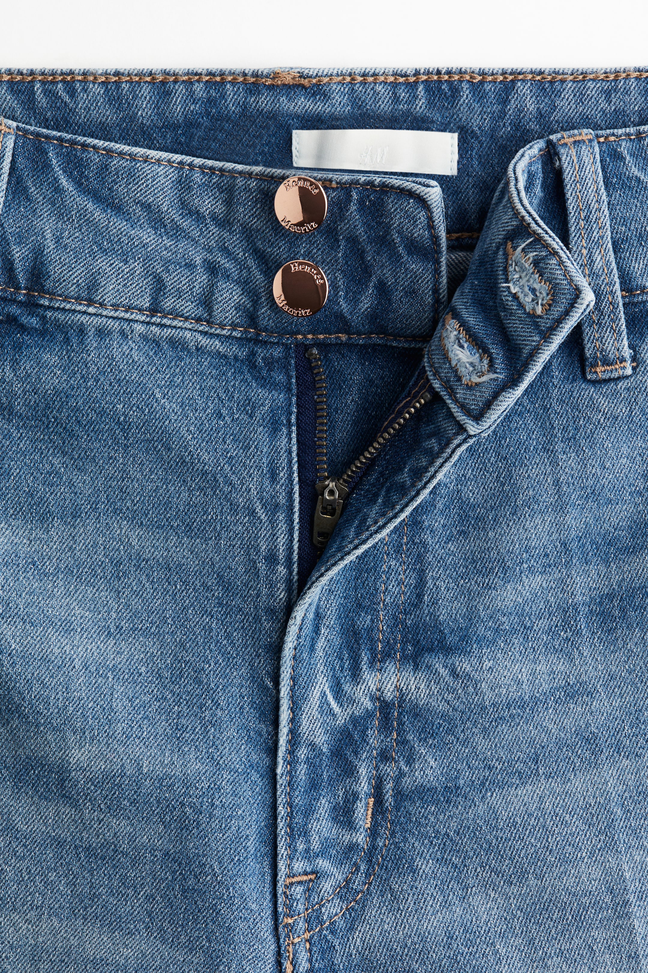 Visualizza immagine più grande: Vista FRONTALE di jeans in denim blu, che mostrano una vita alta con una zip parzialmente aperta e due bottoni rotondi color oro rosa. I bottoni sono incisi con "Hennes Mauritz" e un'etichetta "H&M" è visibile sulla fascia interna della vita.