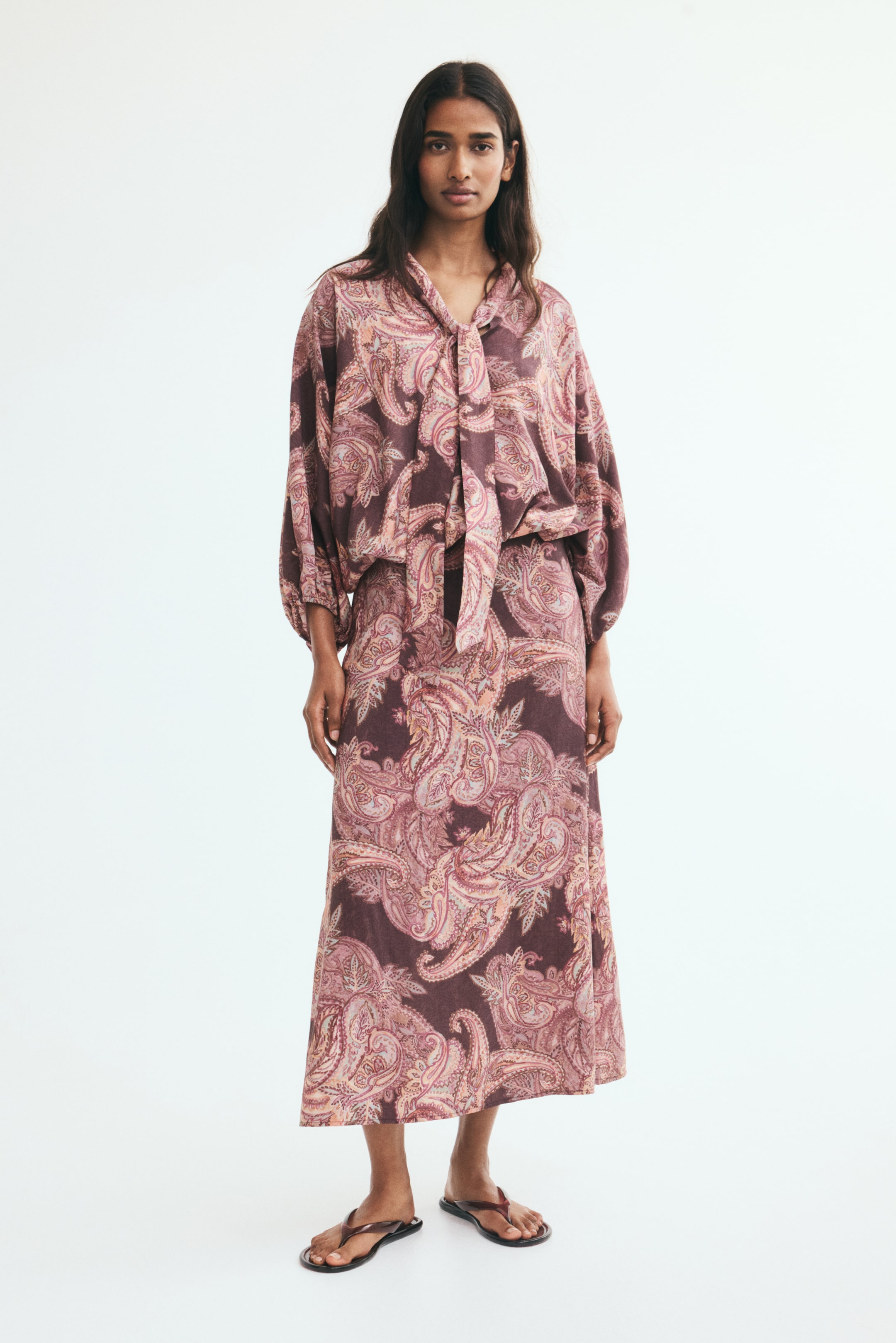 View larger image: The model is wearing a calf-length skirt featuring an all-over purple and pink paisley pattern. The skirt is styled with a matching long-sleeved, tie-neck blouse in the same pattern and a pair of dark flat sandals.