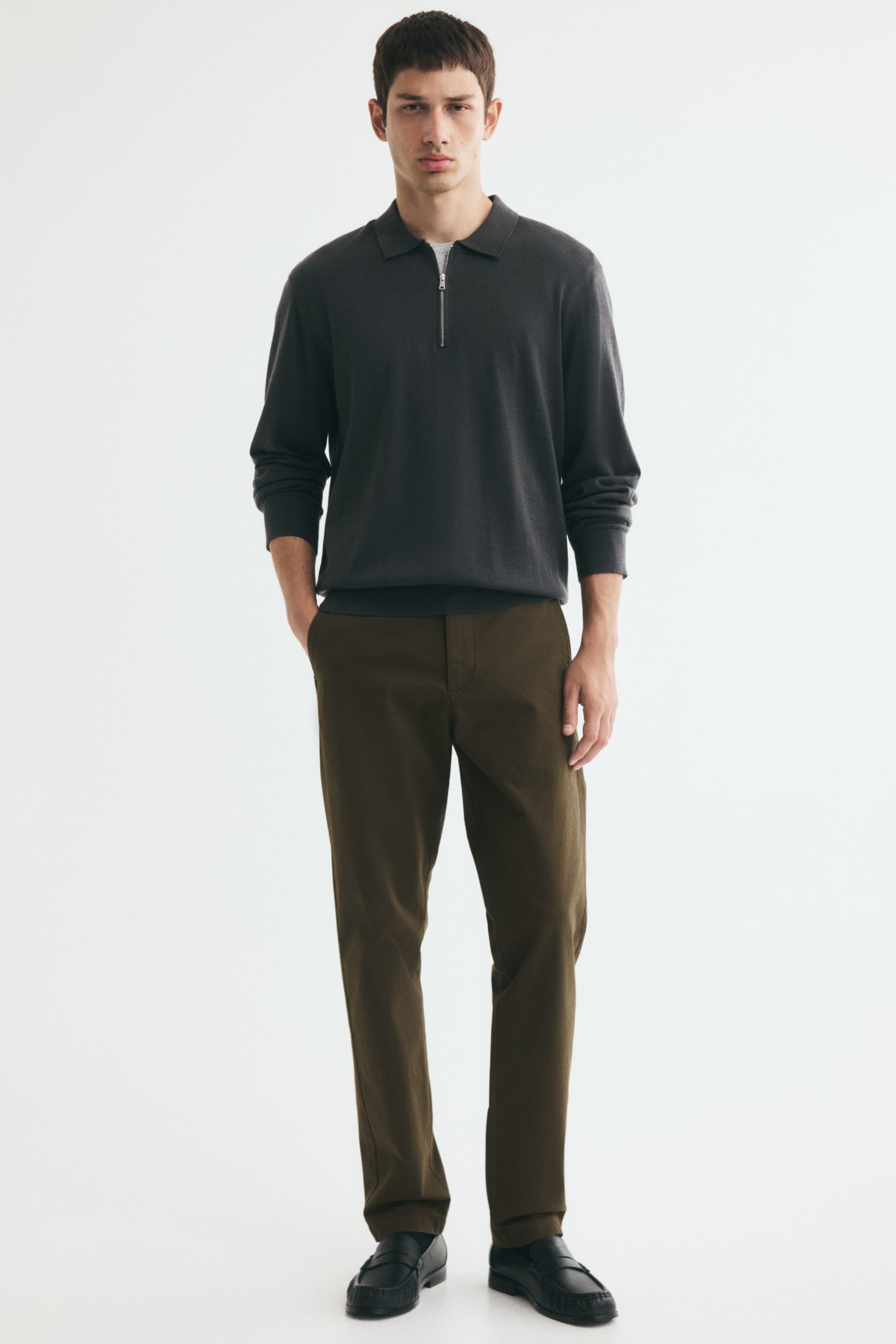 View larger image: Olive brown slim-fit chinos in stretch cotton twill are worn by the model, who is standing with one hand tucked into a diagonal side pocket. The trousers are styled with a charcoal grey long-sleeve half-zip polo shirt and black leather loafers.