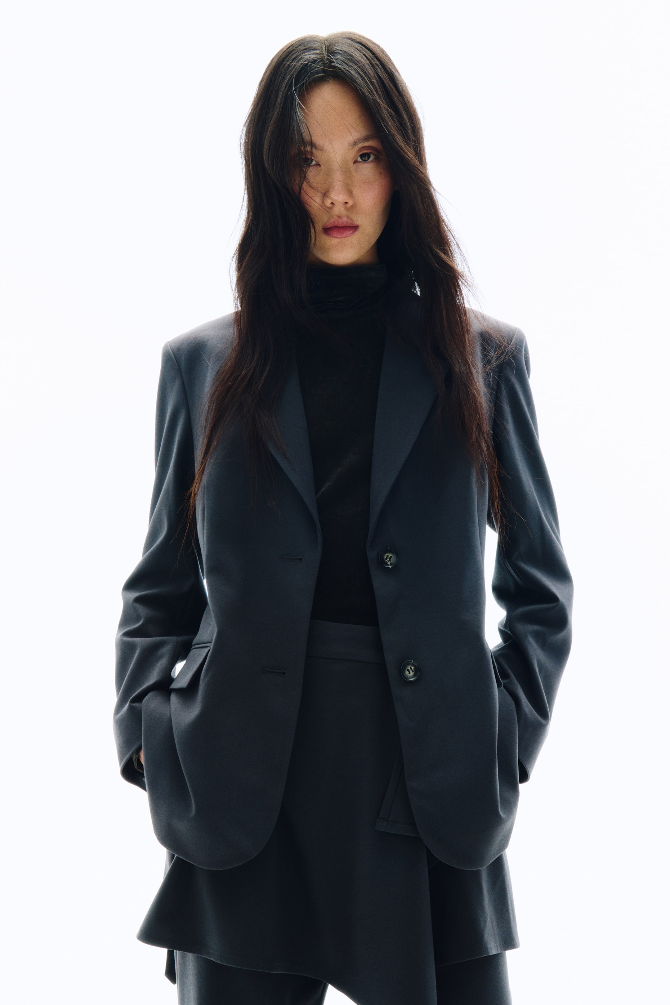 拡大画像を表示: ダークグレーのシングルブレストブレザーに、ノッチドラペル、フロントの2ボタン、フラップポケットを備えた一着を、ブラックのタートルネックの上から着用しています。ボトムスには、アシンメトリーヘムが特徴の同色系ダークグレーのボトムスをコーディネートしています。