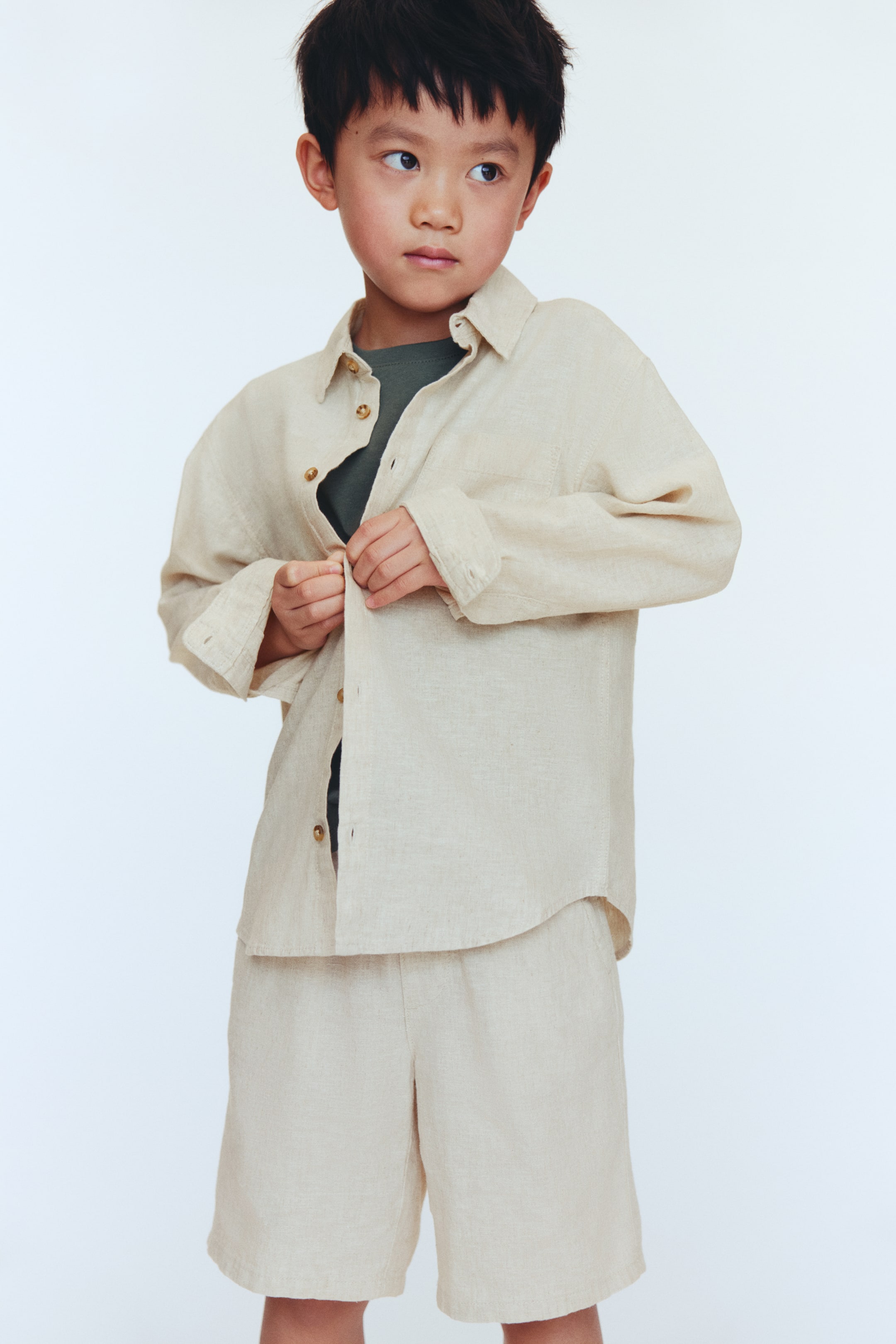 Zobacz większy obraz: Beżowy zestaw lniany składa się z koszuli z długim rękawem i kołnierzykiem, brązowymi guzikami z przodu oraz kieszonką na lewej piersi, noszonej obecnie otwartej na ciemnozielonym t-shircie. Dopasowane, luźne szorty do kolan dopełniają zestaw.