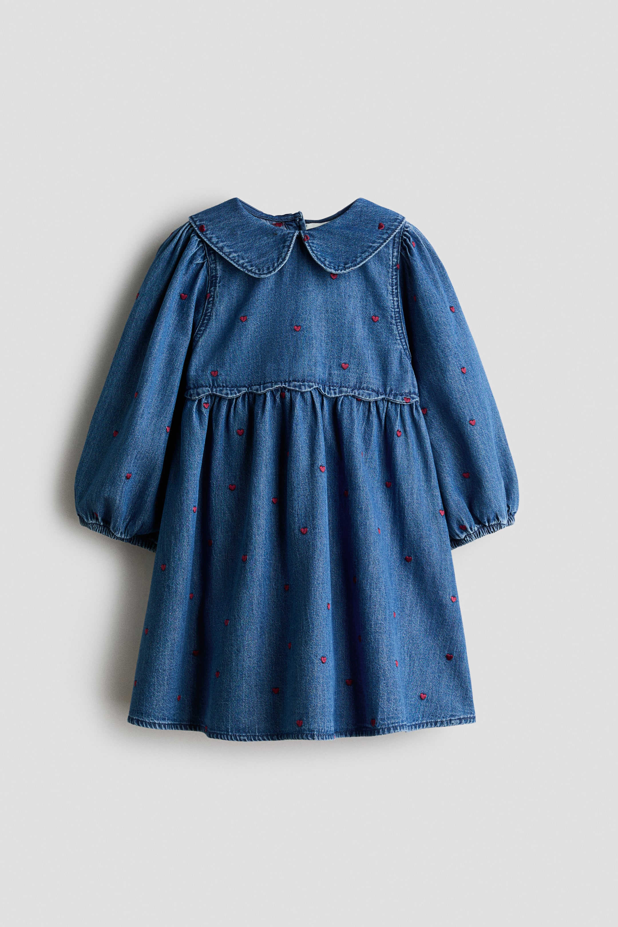 View larger image: A blue cotton denim A-line dress is shown from the front, featuring an all-over pattern of small, embroidered red hearts. The dress displays a rounded collar, long balloon sleeves with narrow elastic cuffs, and a gathered seam above the waist with a small frill trim.