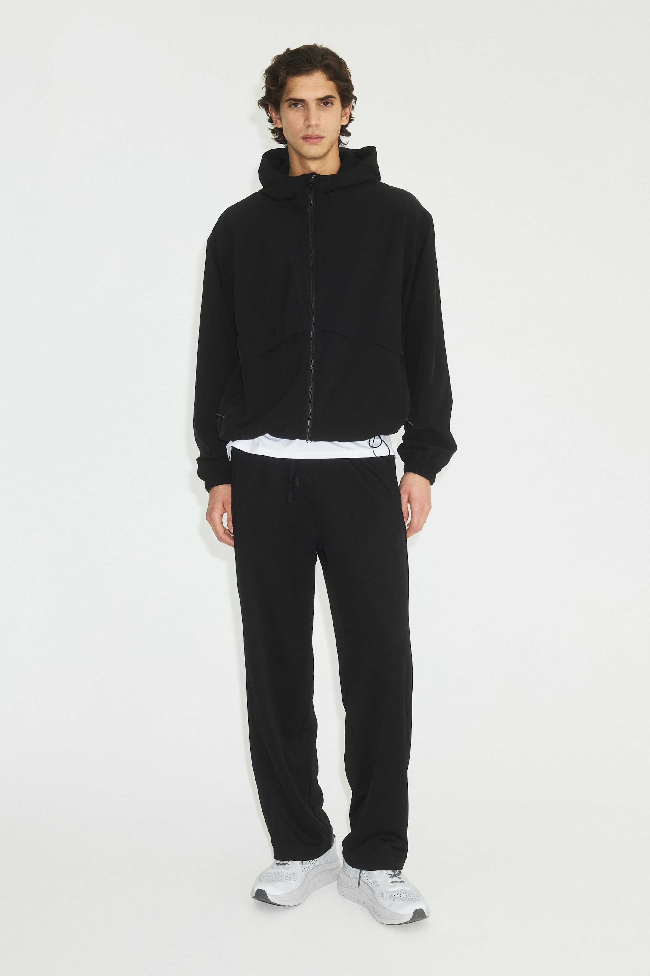 Grotere afbeelding bekijken: Een zwarte hoodie met volledige ritssluiting, naar voren gericht, toont een trekkoordzoom en een capuchon, terwijl er een wit T-shirt onder wordt gedragen. Zwarte sweatpants met rechte pijpen hebben een trekkoord in de taille, en witte en grijze sportsneakers zijn zichtbaar.