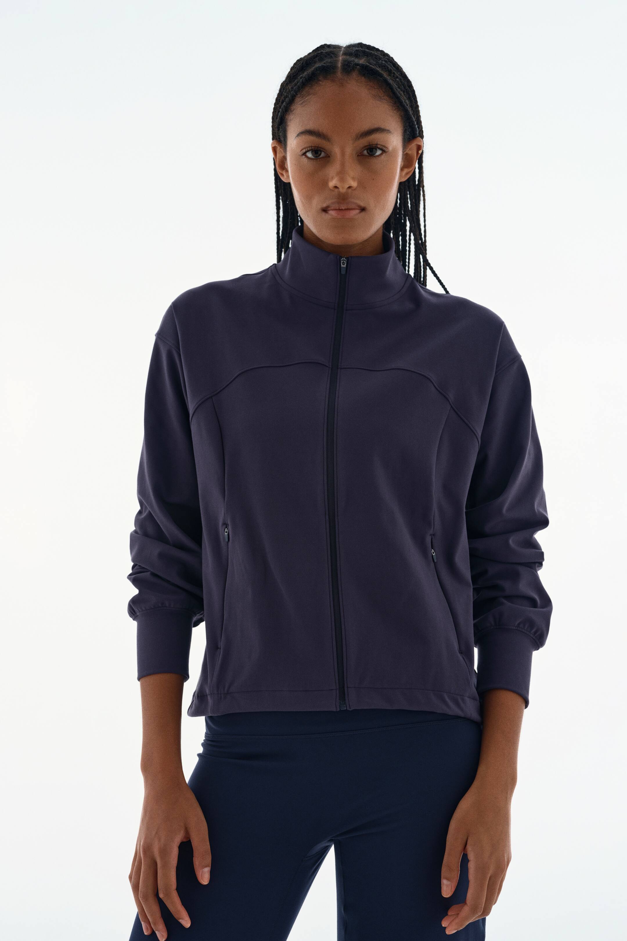 View larger image: A dark purple, full-zip athletic jacket featuring a high collar, curved chest seams, and zippered side pockets is being worn by the model. The jacket is paired with dark blue leggings.