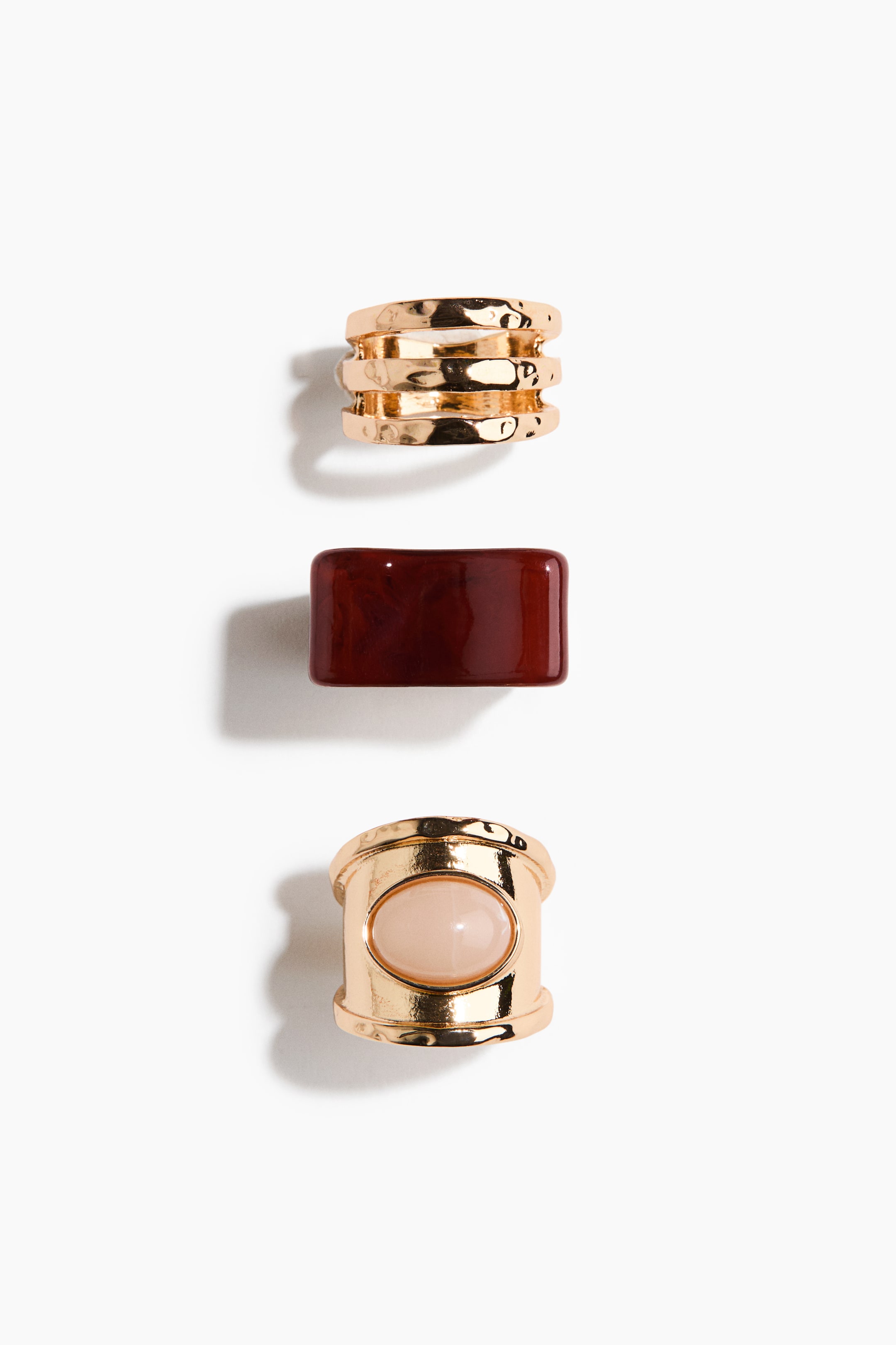 Ver imagen más grande: Un conjunto de tres piezas de joyería distintas se presenta, dispuestas verticalmente. La pieza superior es un anillo ancho de tono dorado con tres bandas martilladas. La pieza del medio es un componente rectangular, translúcido de color rojo oscuro, y la pieza inferior es un anillo ancho de tono dorado que exhibe una piedra cabujón ovalada de color rosa claro en el centro.