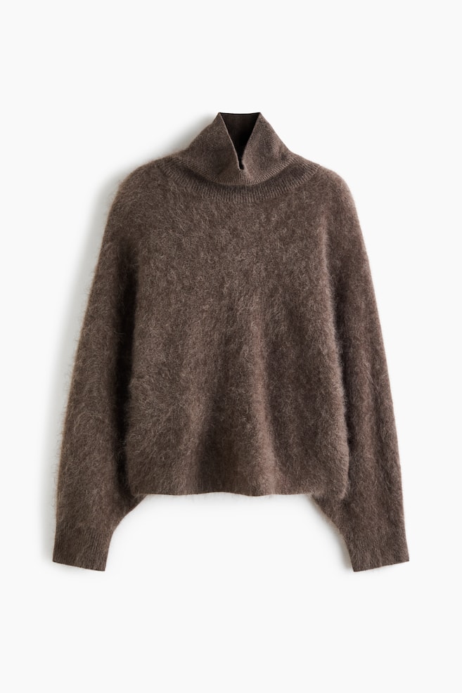 An oversized brown jumper in a brushed wool knit is shown from a front angle, highlighting its soft, fuzzy texture. The jumper is featuring a high, ribbed turtleneck and long, voluminous sleeves ending in ribbed cuffs. - 1