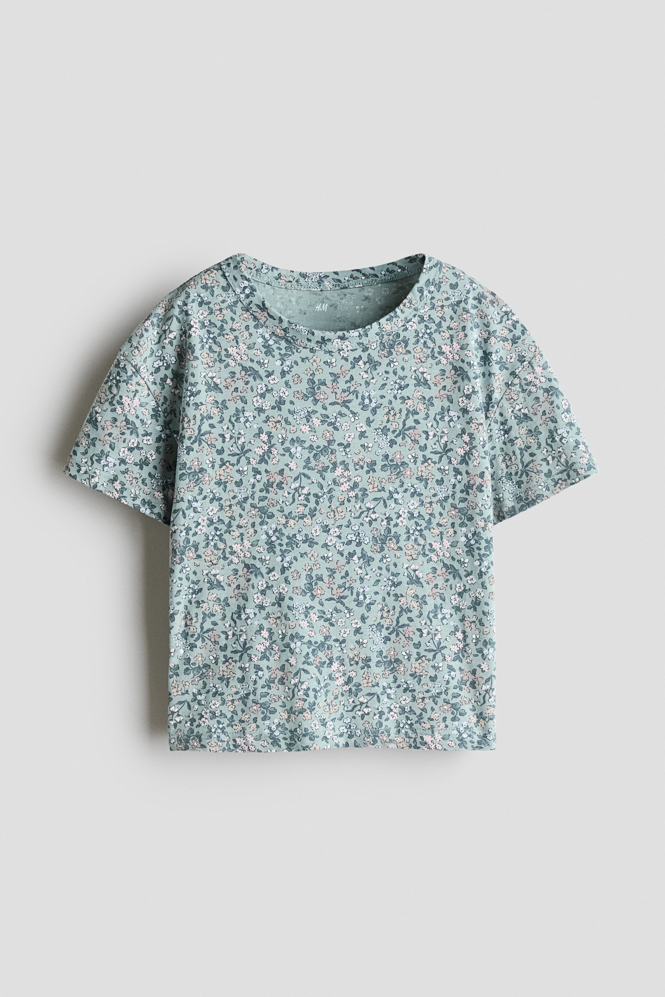 Xem ảnh lớn hơn: Áo thun hướng mặt trước bằng vải jersey cotton màu xanh lá nhạt được in họa tiết hoa nhỏ màu trắng và hồng phủ toàn bộ bề mặt. Áo có cổ tròn bo gân, vai trễ và tay ngắn.
