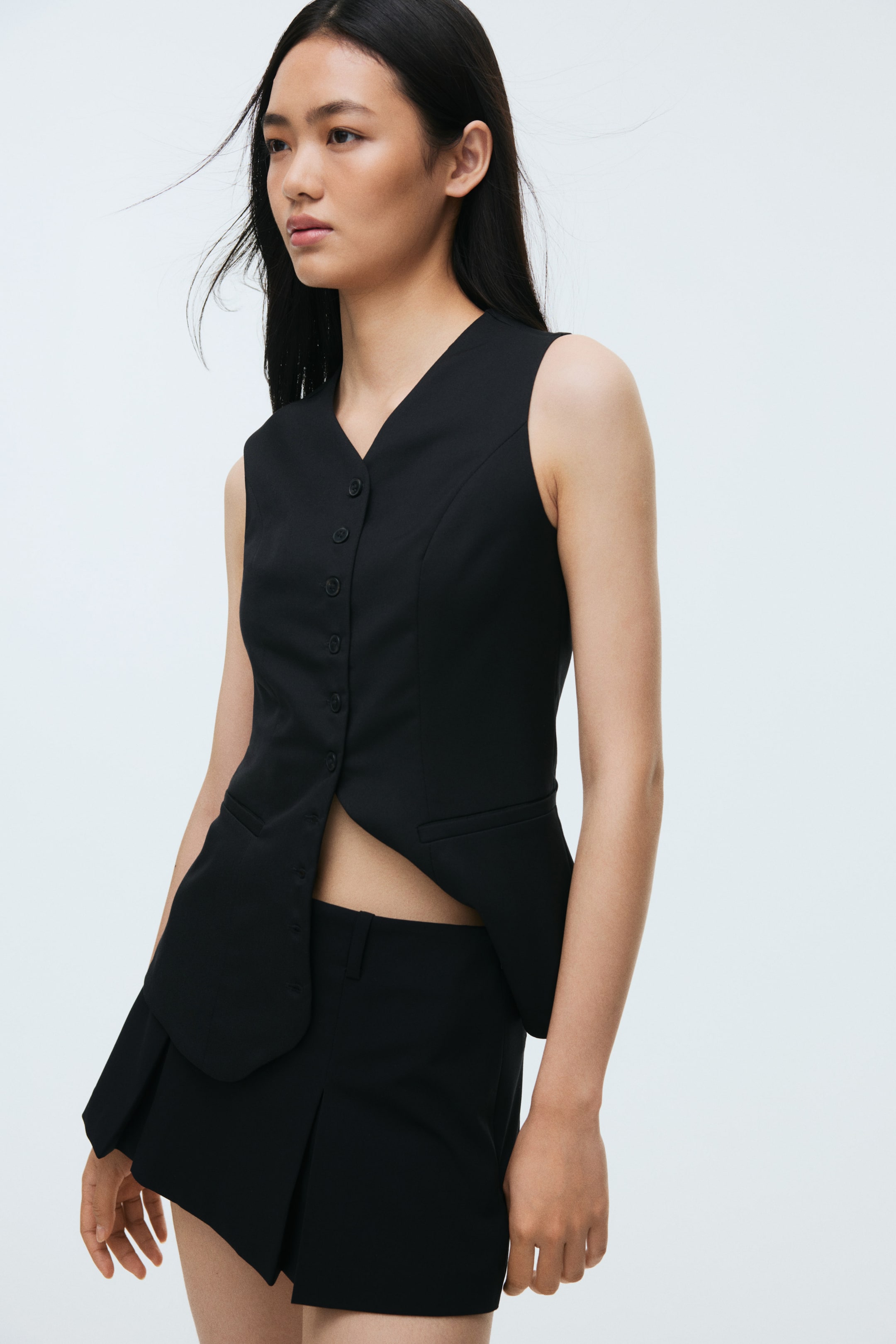 Ampliar la imagen: Un chaleco negro sin mangas y unos shorts plisados a juego se presentan desde un ángulo de tres cuartos. El chaleco cuenta con un escote en V, un cierre de botones en la parte frontal y dos bolsillos delanteros con ribete, con un dobladillo asimétrico que se curva hacia afuera y revela el abdomen; los shorts plisados presentan un corte corto y pliegues frontales.
