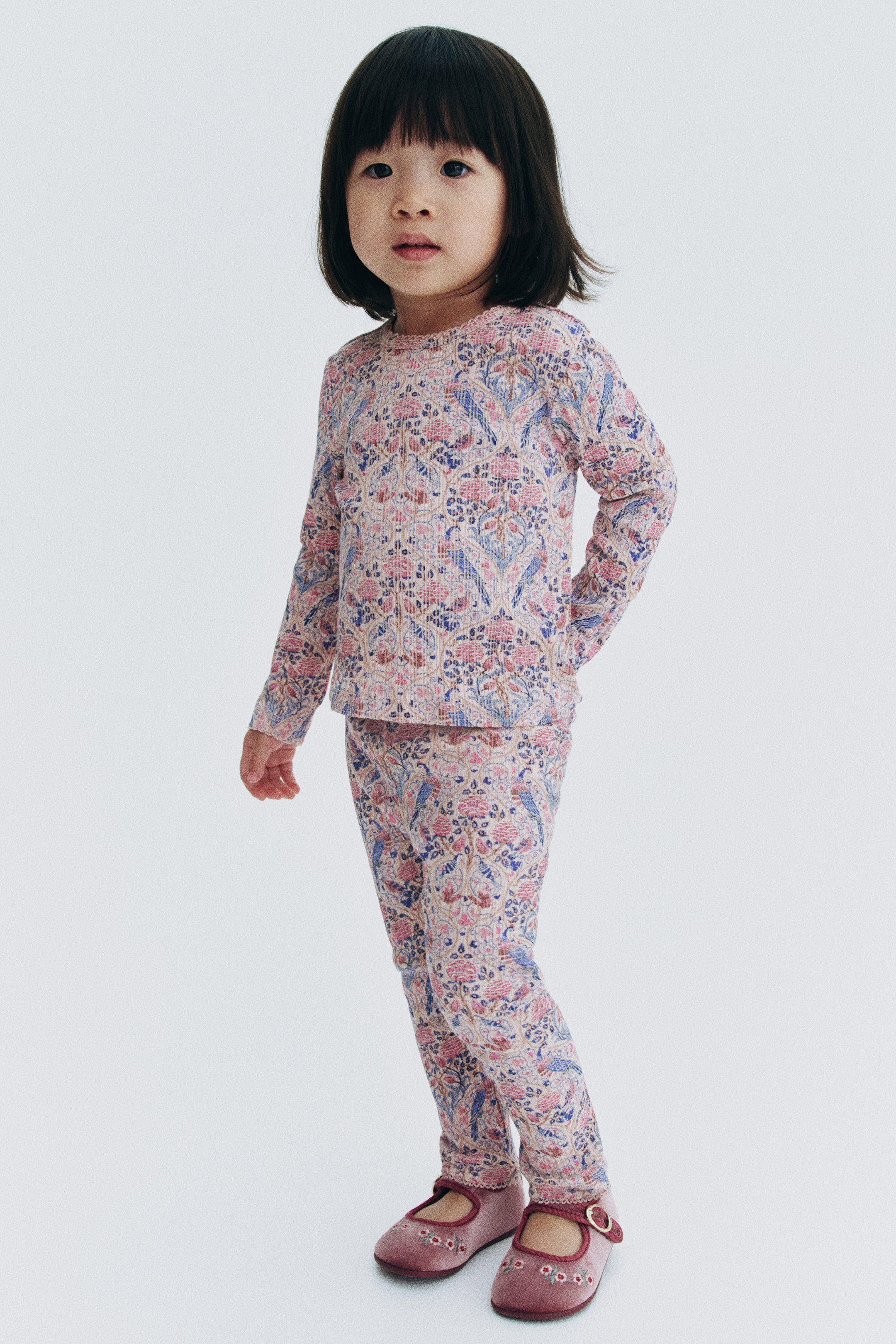 Vis større bilde: Et matchende sett med langermet topp og leggings vises i en trekvart frontvisning, med et heldekkende blomsterprint i rosa og blått med ornamentale detaljer på ribbestrikket bomullsjersey. Toppen har rund hals med en delikat picot-kant, mens de matchende slim fit-leggingsene smalner ned mot anklene og er kombinert med rosa Mary Jane-sko i fløyel.