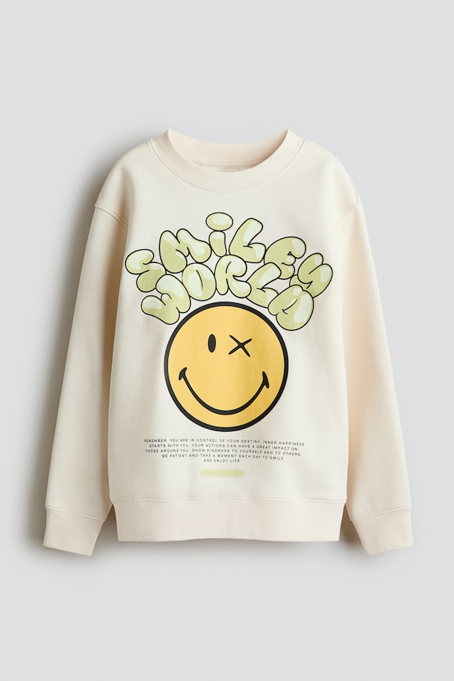 A cream lightweight sweatshirt is facing front, featuring a round, rib-trimmed neckline, dropped shoulders, and long sleeves with ribbed cuffs and hem. The front is displaying print motifs, including a large yellow smiley face with an X-eye, bubbly green "smile world" text above it, and smaller black text below. - 1