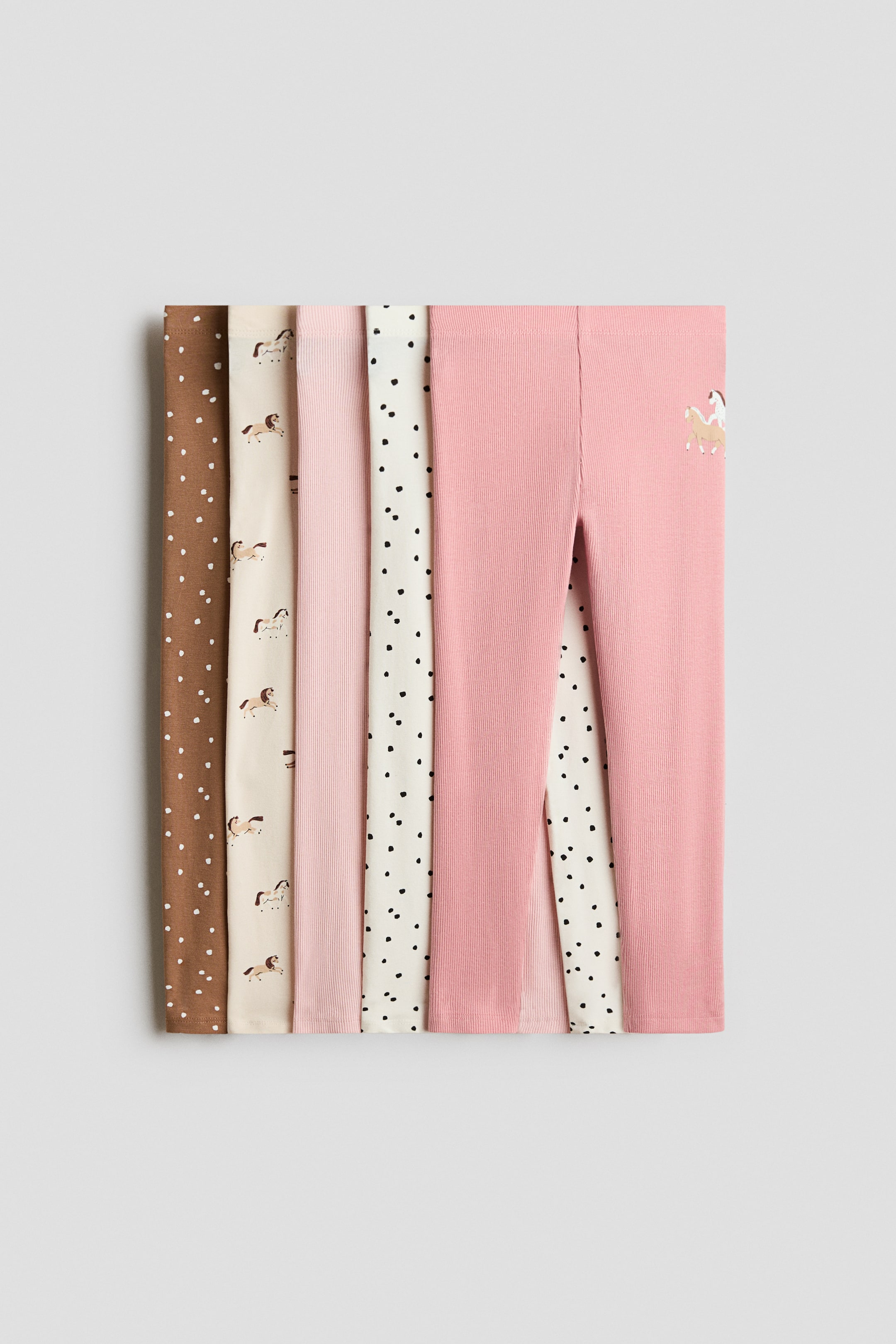Voir une image plus grande: Une pile verticale de leggings en jersey de coton doux avec taille élastique est présentée, montrant leur devant. Cinq paires différentes mettent en valeur divers motifs, dont un modèle brun à pois blancs, un modèle crème avec motifs de chevaux, un modèle rose pâle côtelé, un modèle crème avec motifs de cœurs noirs et un modèle rose côtelé avec un petit cheval brodé.