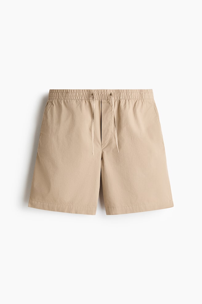 Beige twill shorts are presented lying flat, facing forward, showcasing a relaxed silhouette. They feature an elasticated drawstring waist with visible drawstrings, a fake fly, and diagonal side pockets. - 1