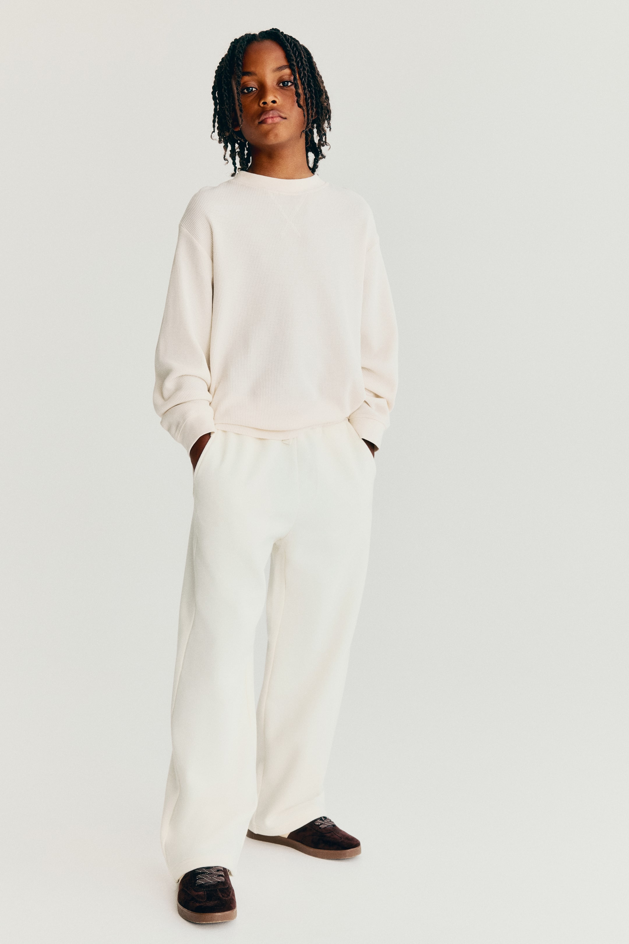 View larger image: Off-white track pants are shown from the front, featuring a relaxed, wide-leg silhouette and diagonal side pockets. The pants are styled with a matching crewneck sweatshirt and dark brown sneakers.