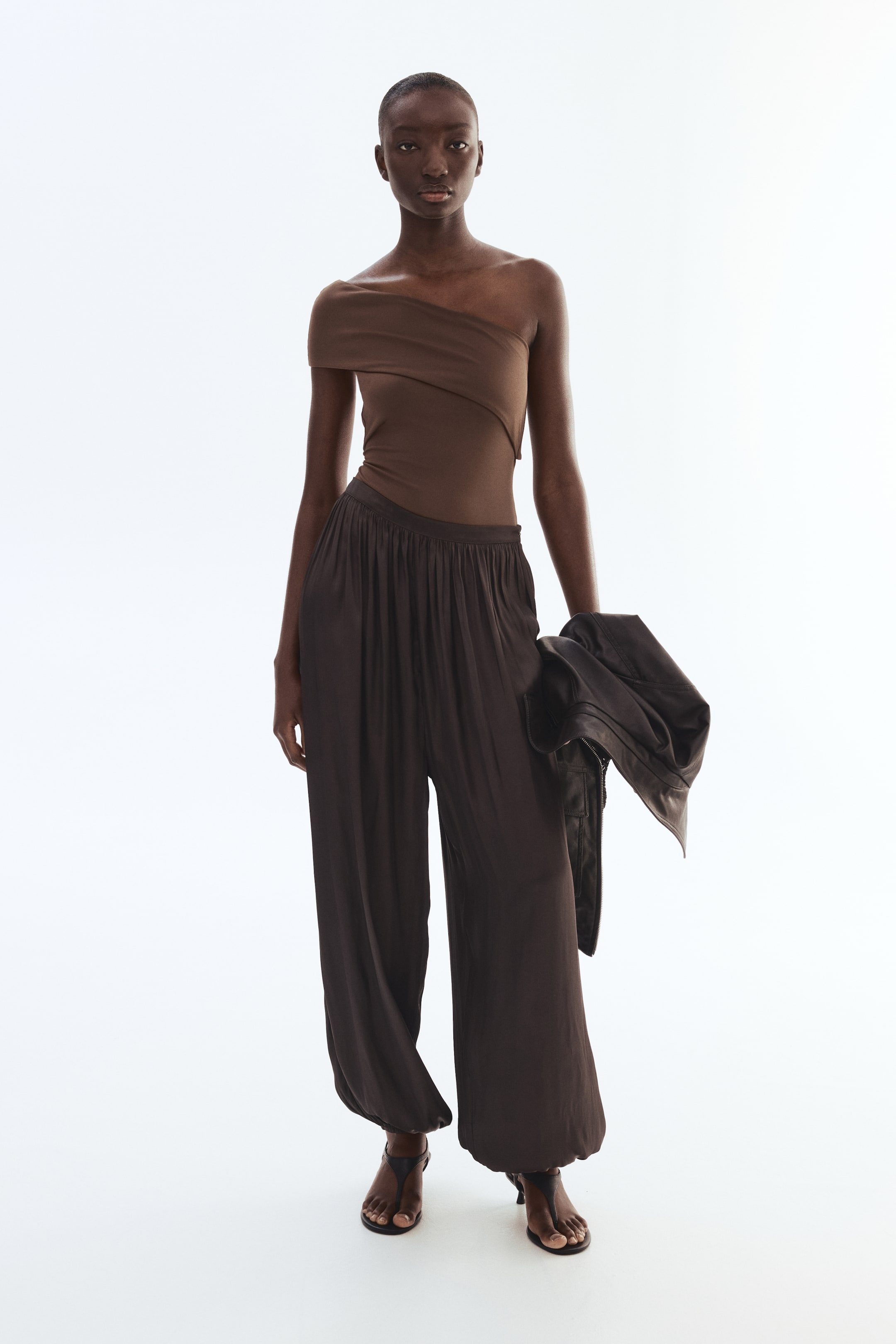 View larger image: Dark brown, loose-fitting pants are showcased from a front-facing perspective, featuring a gathered high waist and wide legs that taper to elasticated hems, creating a voluminous silhouette.