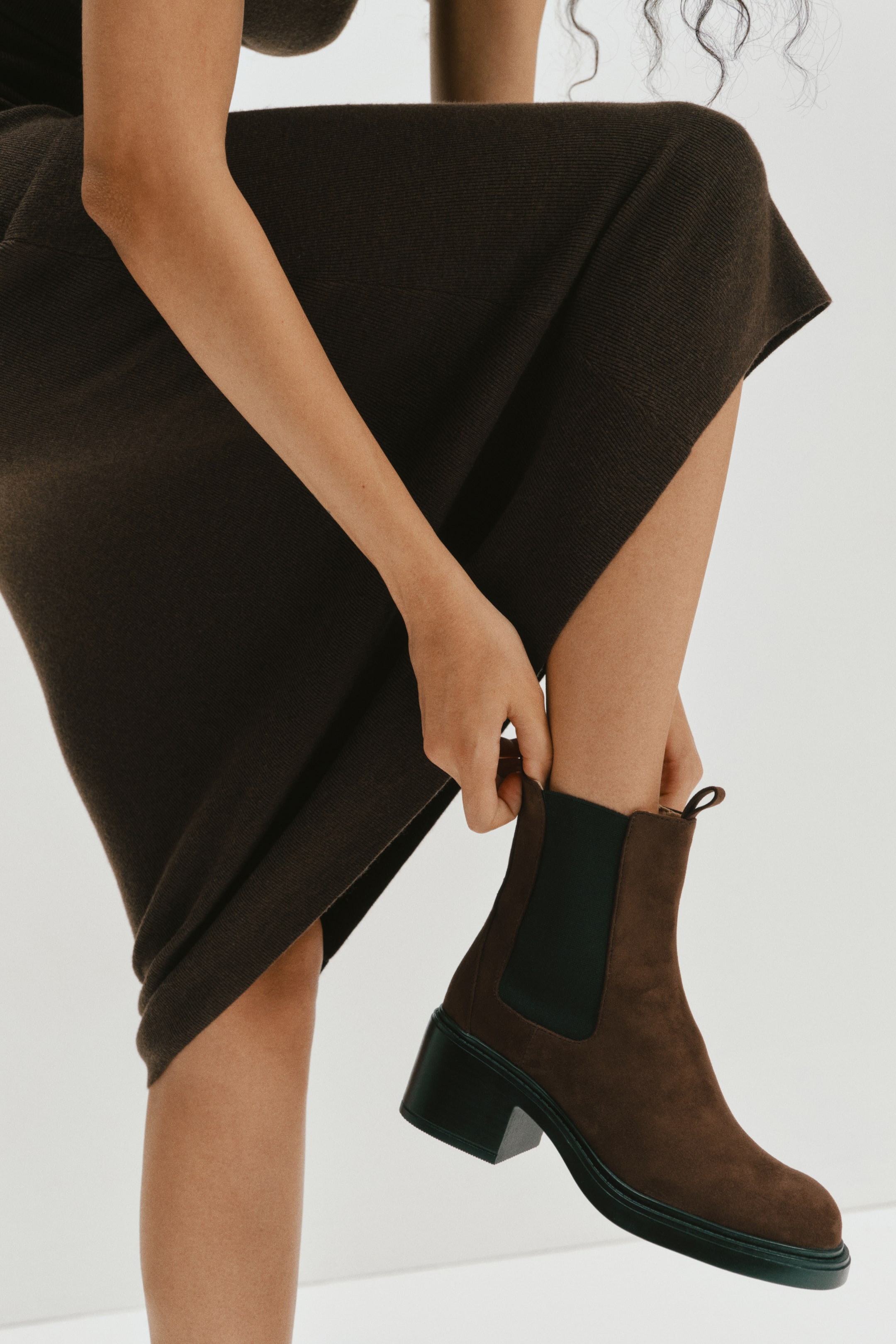 Ver imagen más grande: La modelo lleva unas botas Chelsea de ante marrón con punteras redondeadas, tacones negros de bloque y paneles laterales elásticos negros. La modelo está tirando del tirador trasero de una de las botas mientras lleva una falda de punto marrón oscuro.