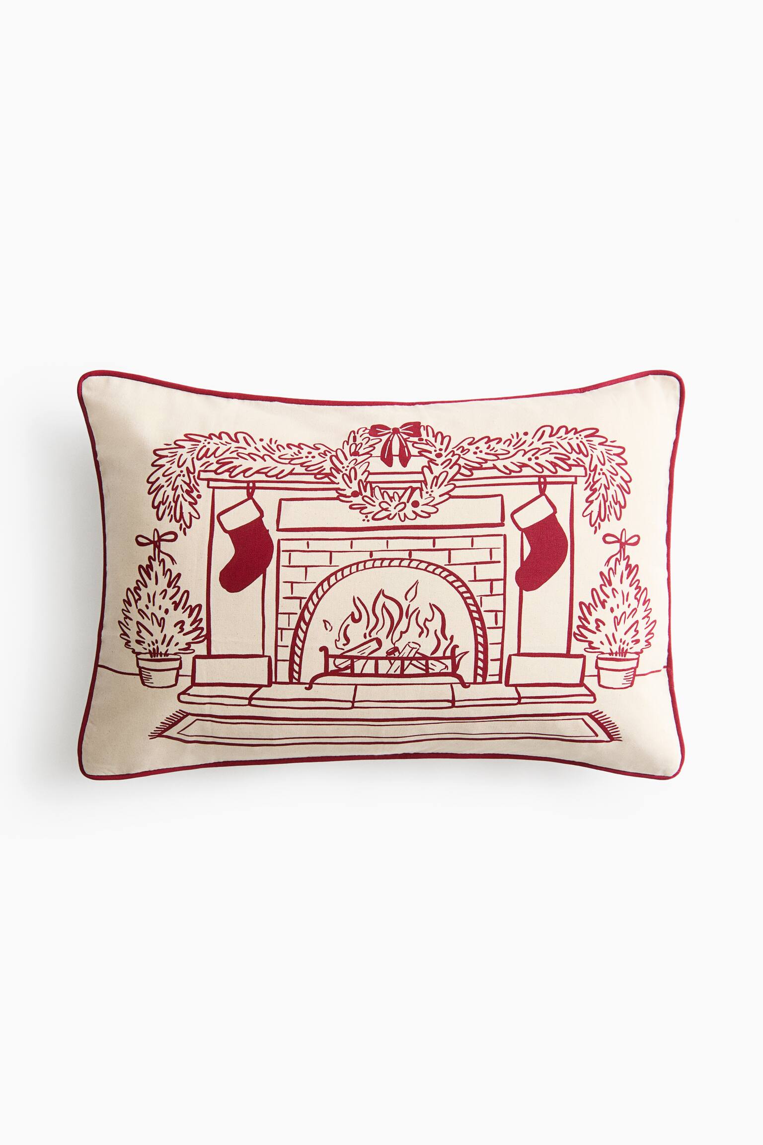 A rectangular off-white lumbar pillow is shown from the front, featuring a red line-drawn illustration of a cosy fireplace scene. The illustration shows a burning fireplace adorned with a mantel garland, a central wreath, hanging stockings, and potted evergreen trees on either side, all framed by red piped trim along the edges.
