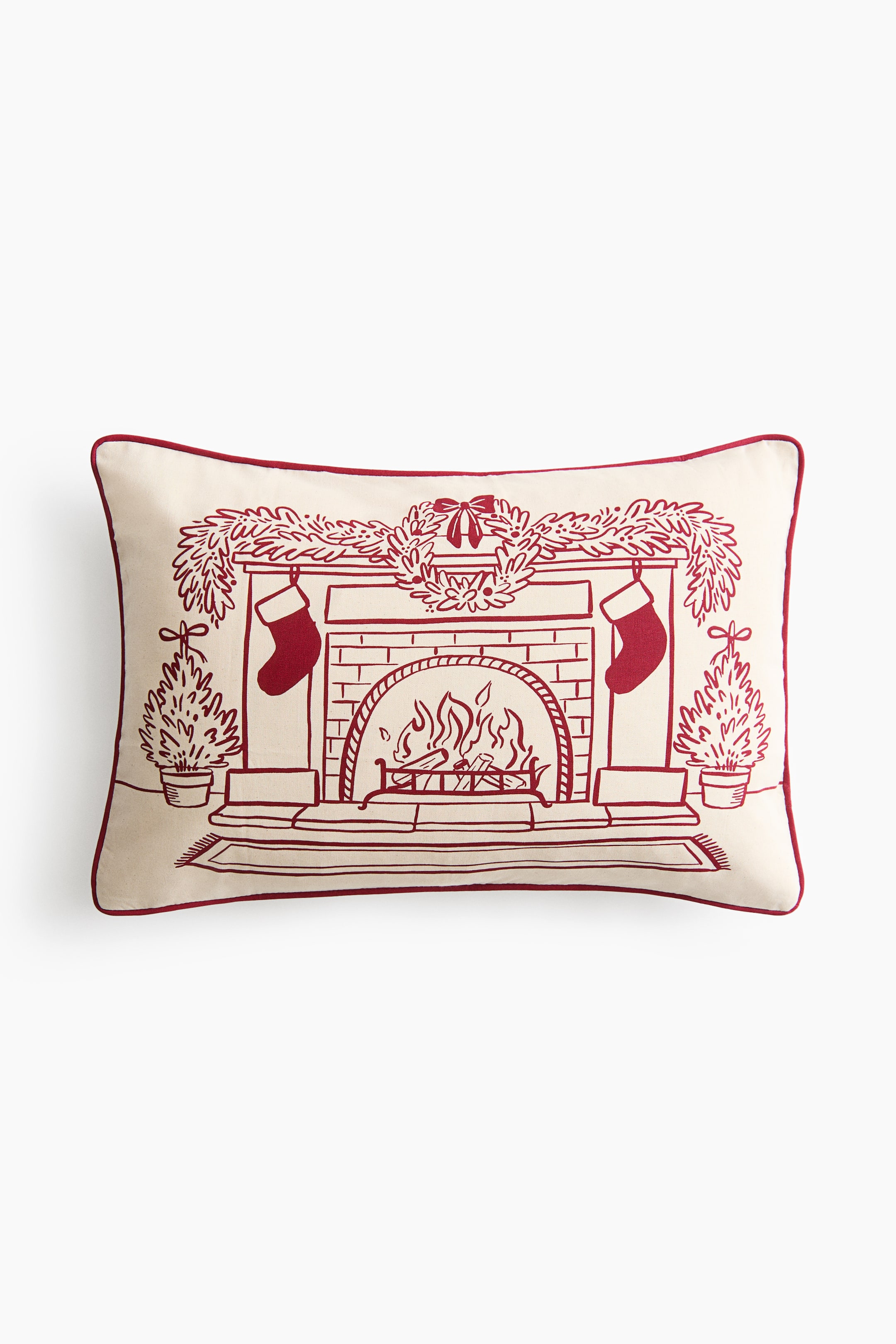 View larger image: A rectangular off-white lumbar pillow is shown from the front, featuring a red line-drawn illustration of a cosy fireplace scene. The illustration shows a burning fireplace adorned with a mantel garland, a central wreath, hanging stockings, and potted evergreen trees on either side, all framed by red piped trim along the edges.