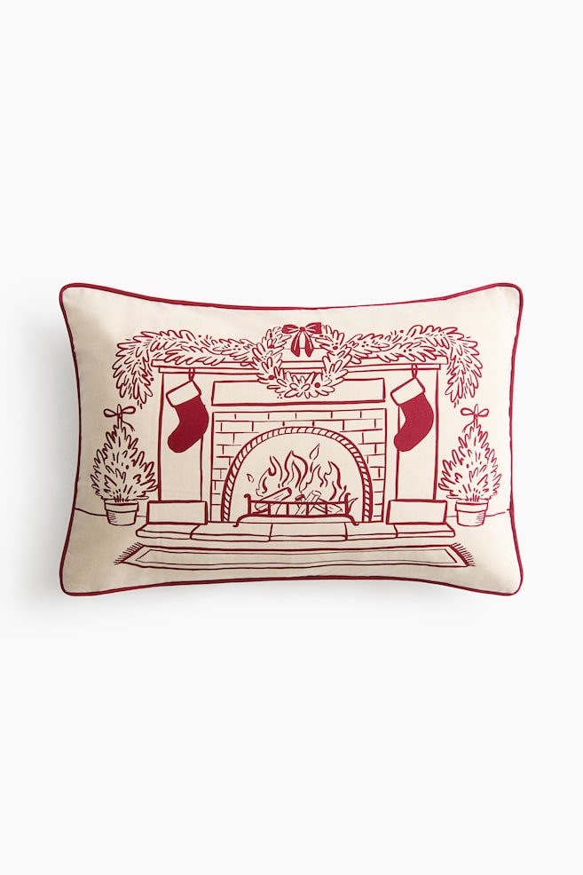 En rektangulÃ¤r benvit lÃ¤ndkudde Ã¤r vÃ¤nd framÃ¥t och visar en rÃ¶d linjetecknad illustration av en mysig spisÃ¶versikt. Illustrationen fÃ¶restÃ¤ller en brinnande eldstad prydd med en girlang pÃ¥ spiselkransen, en krans i mitten, upphÃ¤ngda strumpor och vintergrÃ¶na trÃ¤d i krukor pÃ¥ vardera sidan, allt inramat av rÃ¶d passpoal lÃ¤ngs kanterna. - 1
