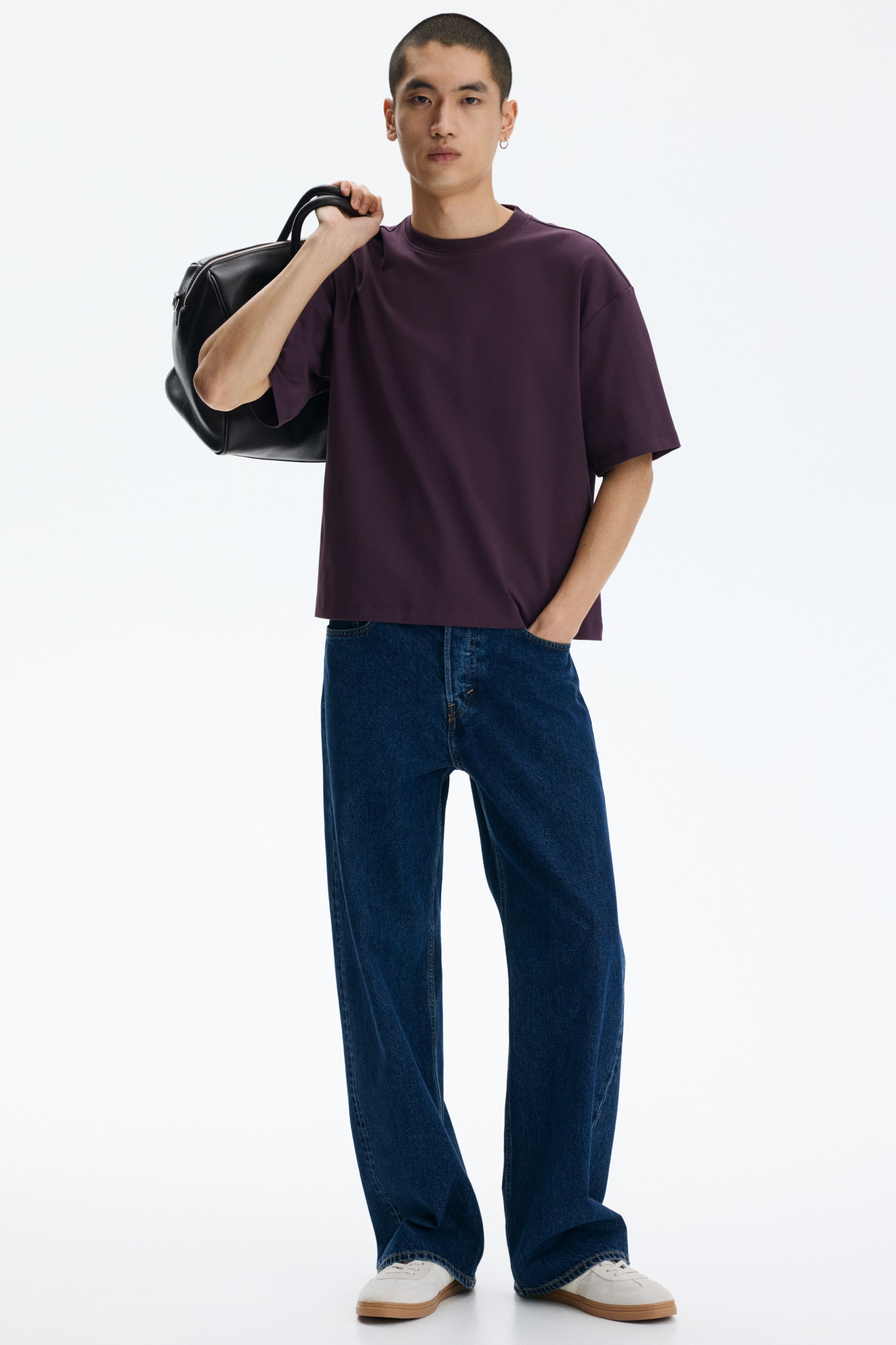 查看大圖: 一件深紫色的圓領T恤正在展示，特點是短而寬的袖子和寬鬆的方形剪裁。它未塞進深藍色牛仔褲中，黑色包帶部分遮住了左肩。