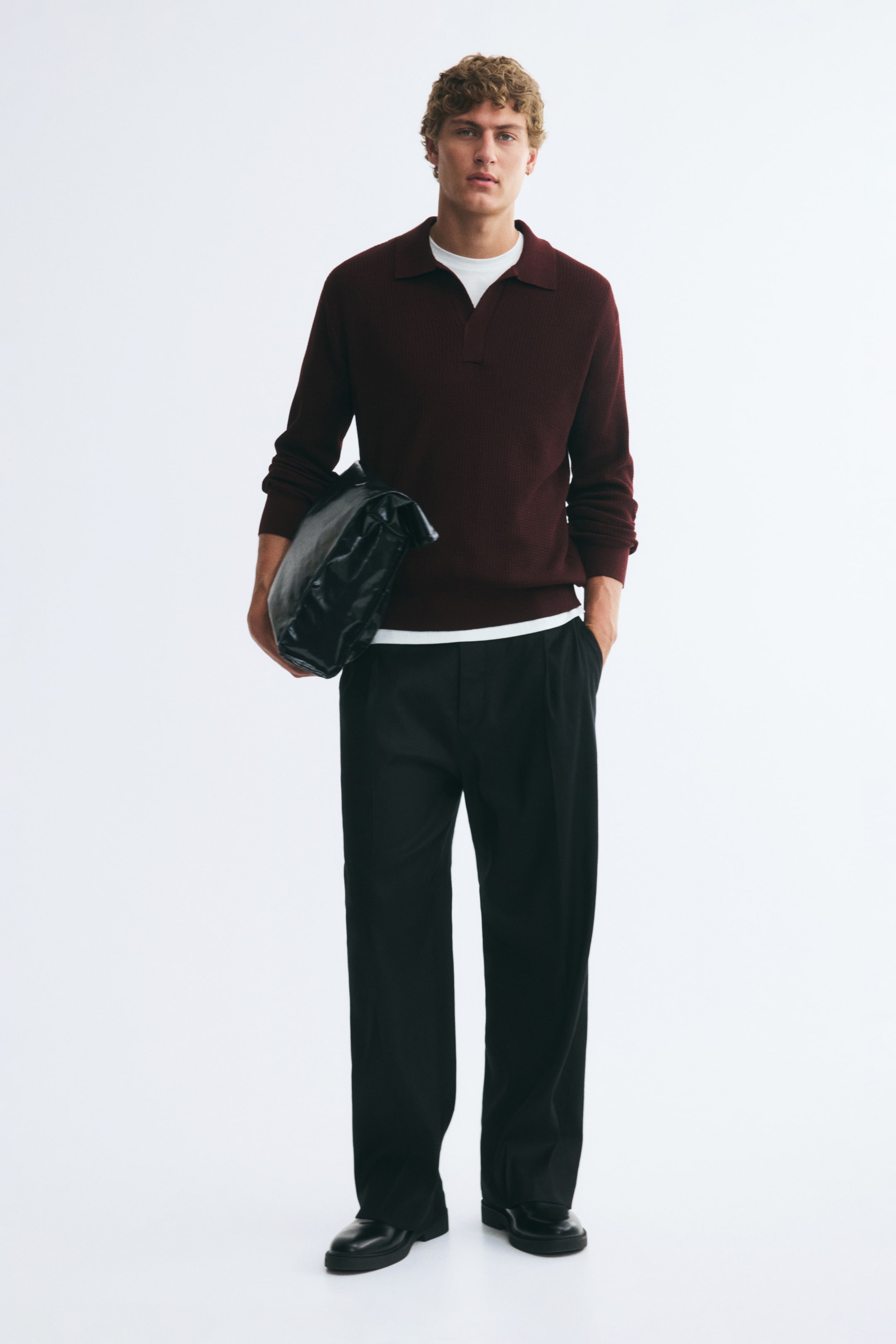 View larger image: A burgundy textured-knit polo shirt with an open collar and long sleeves is being worn by the model, layered over a white T-shirt. The relaxed-fit shirt features ribbed cuffs and hem, and is styled with black trousers while the model is holding a black bag under one arm.