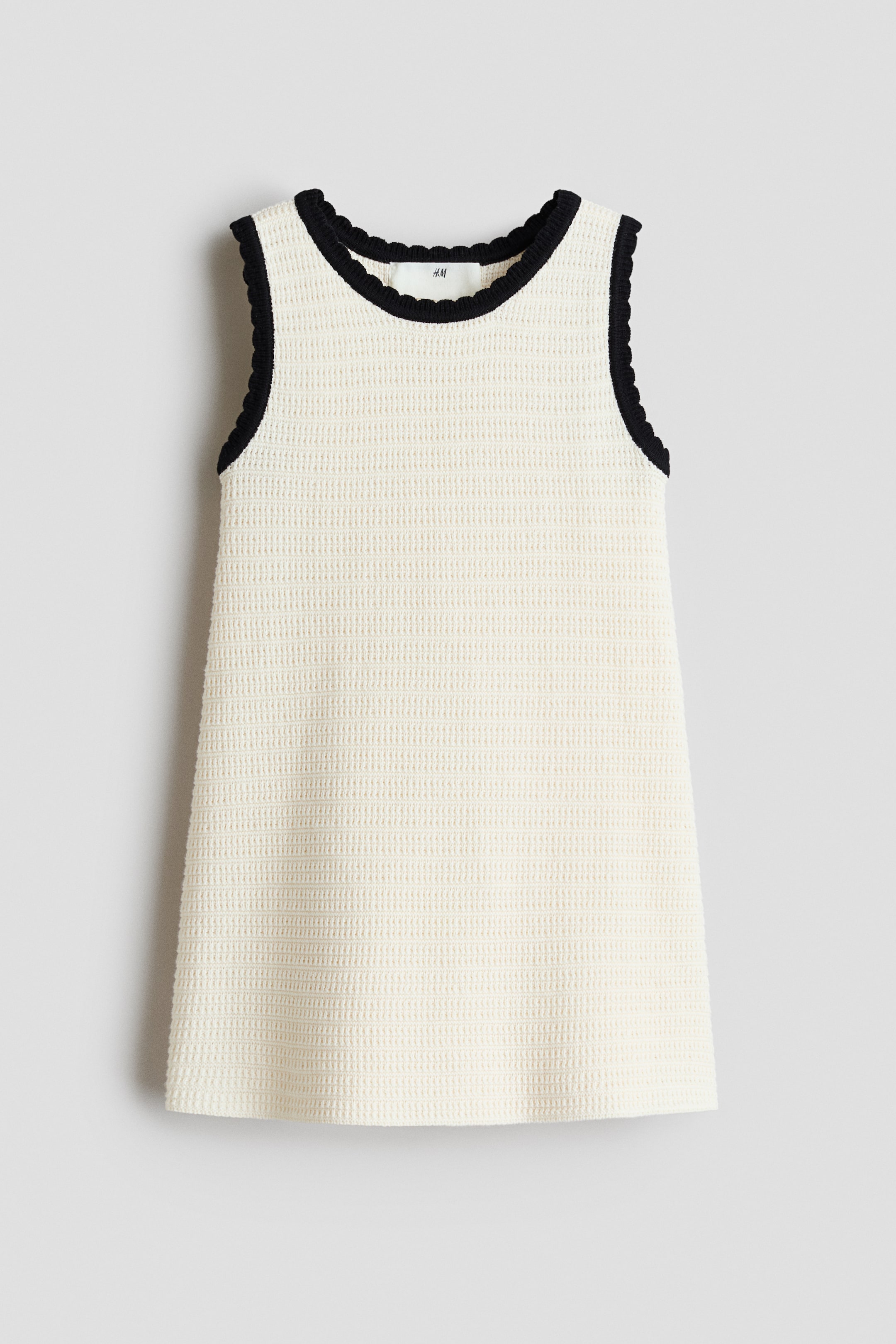 Agrandir l'image: Une robe trapèze sans manches de couleur crème est présentée de face, mettant en avant un tricot texturé. Un bord côtelé noir, festonné, souligne l'encolure ronde et les emmanchures.