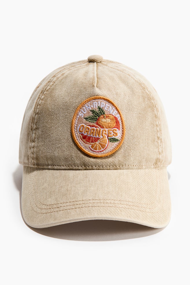 Une casquette de baseball beige présente son devant, arborant un patch brodé circulaire mettant en scène des oranges et des feuilles, avec le texte "oranges mûries au soleil" cousu en blanc. La casquette est conçue avec une visière incurvée et un bouton supérieur recouvert de tissu. - 1