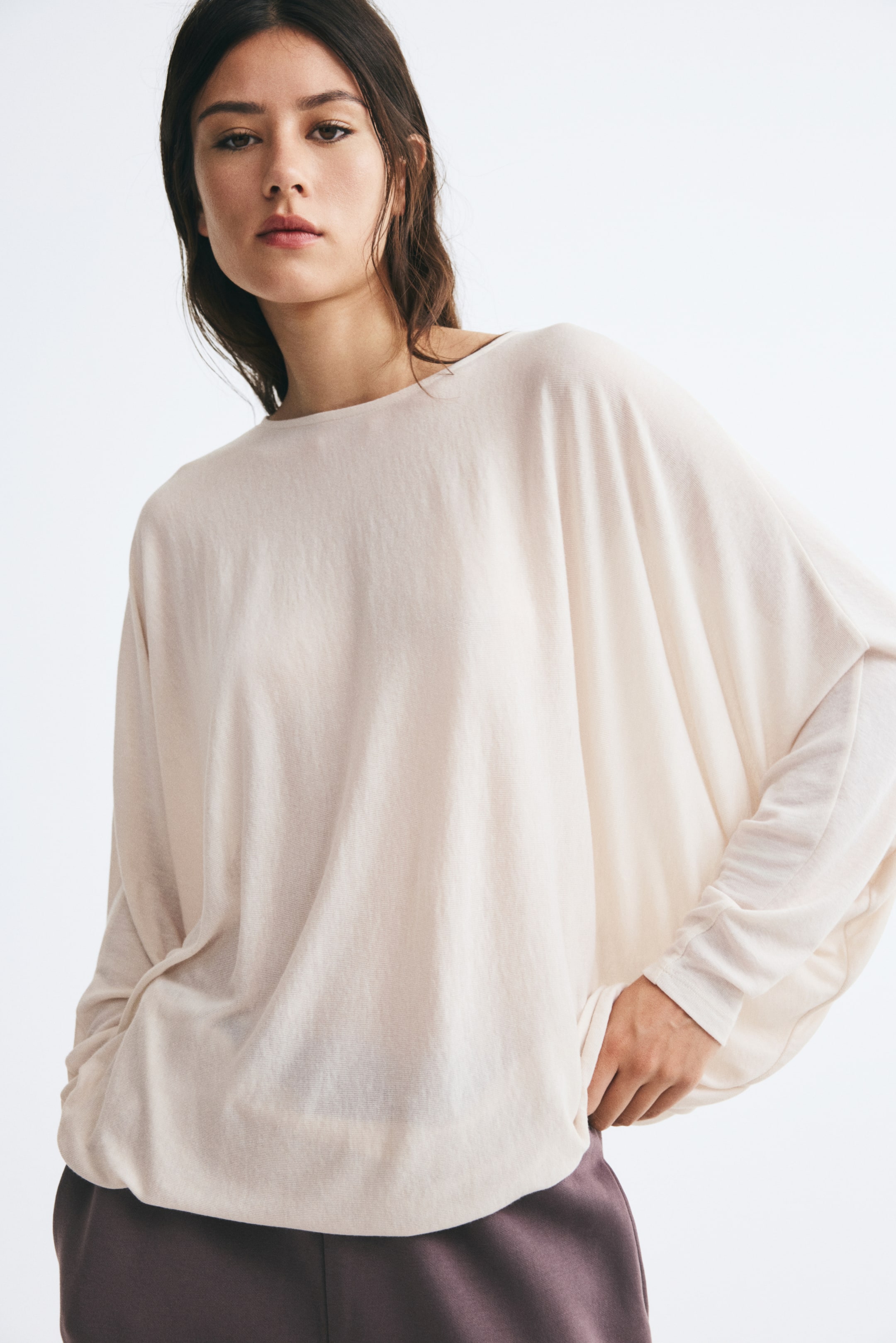View larger image: A loose-fitting, cream-coloured top in a slightly sheer, fine-ribbed jersey is being worn by the model. The top features a round neckline and long, draped batwing sleeves, and is styled over dark purple trousers.