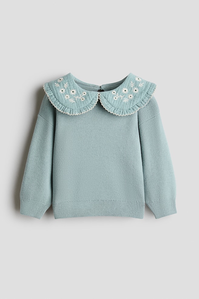 A light blue-green knitted sweater is shown from the front, featuring a large, rounded Peter Pan collar adorned with white embroidered flowers and a ruffled, scalloped trim. The sweater is designed with long sleeves, ribbed cuffs, and a matching ribbed hem. - 1