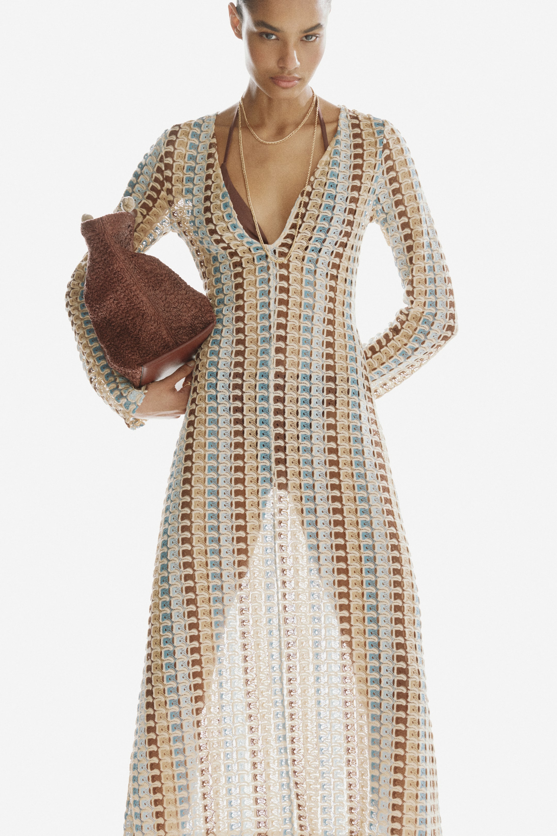 View larger image: The model is wearing a long-sleeved maxi dress crafted from a multi-toned, checkered crochet-look knit. It features a deep V-neckline with a keyhole opening and a hook-and-eye closure at the front. Underneath, a brown bralette and two gold-tone necklaces are visible. The model is also holding a brown textured bag.