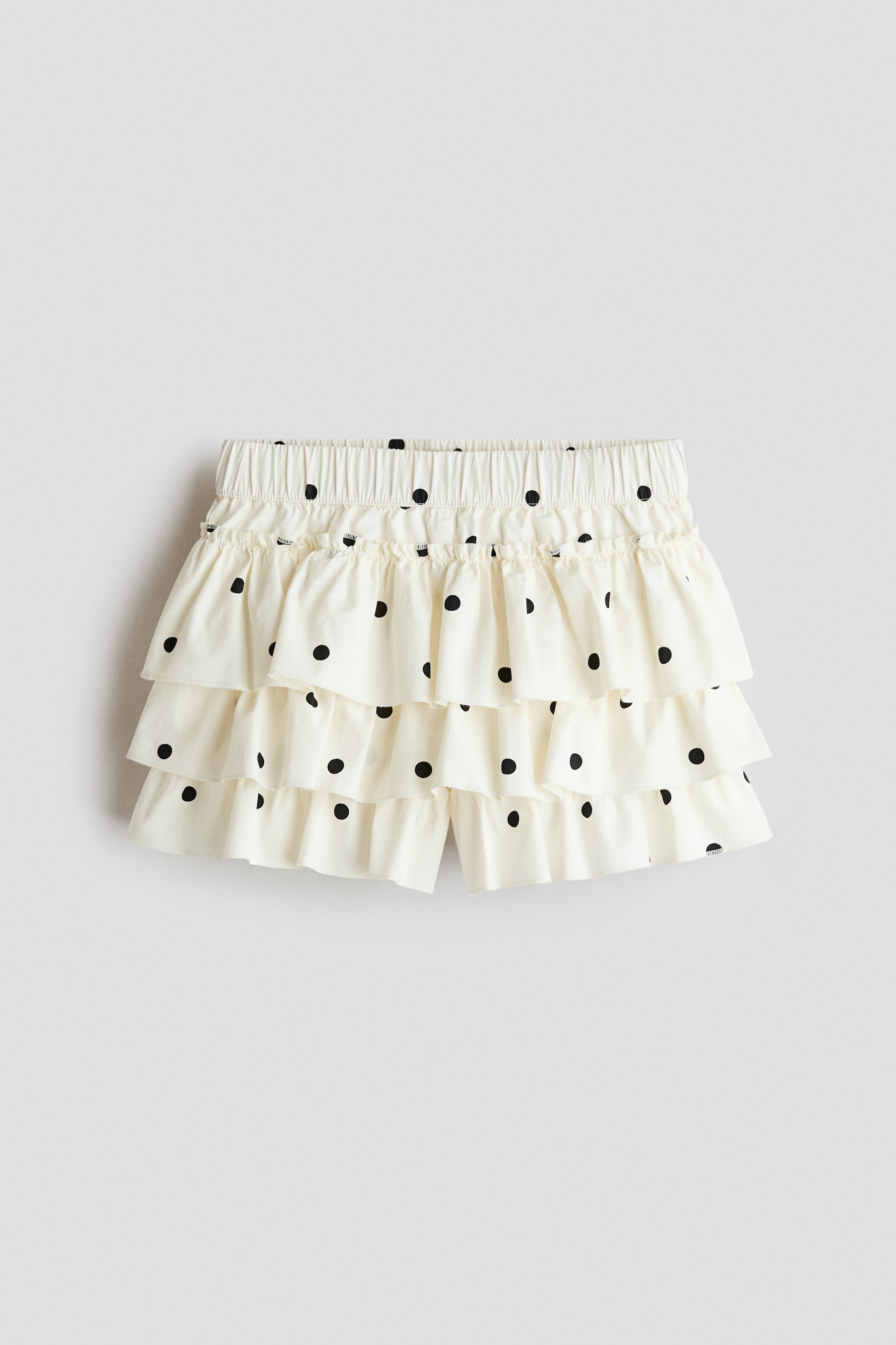 View larger image: A tiered skort in cream-colored cotton seersucker with black polka dots is shown from the front, featuring an elasticated waistband with a small frill trim at the top. The garment features three cascading, ruffled tiers that partially reveal the underlying shorts at the bottom hem.