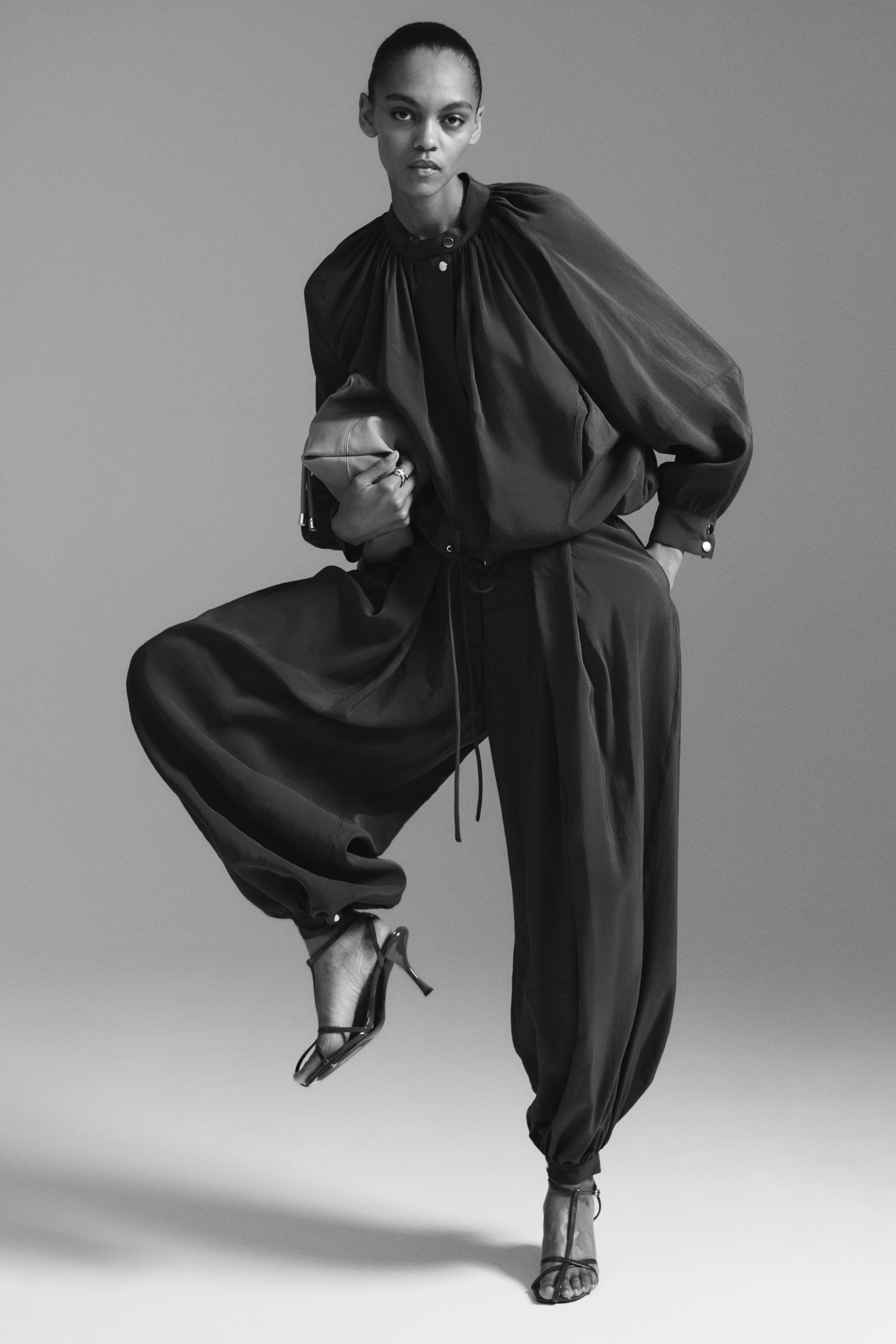 View larger image: The model is wearing loose-fit trousers, characterised by front pleats, balloon legs, and gathered, buttoned cuffs at the hems. The model is posing with one hand resting in a trouser pocket.